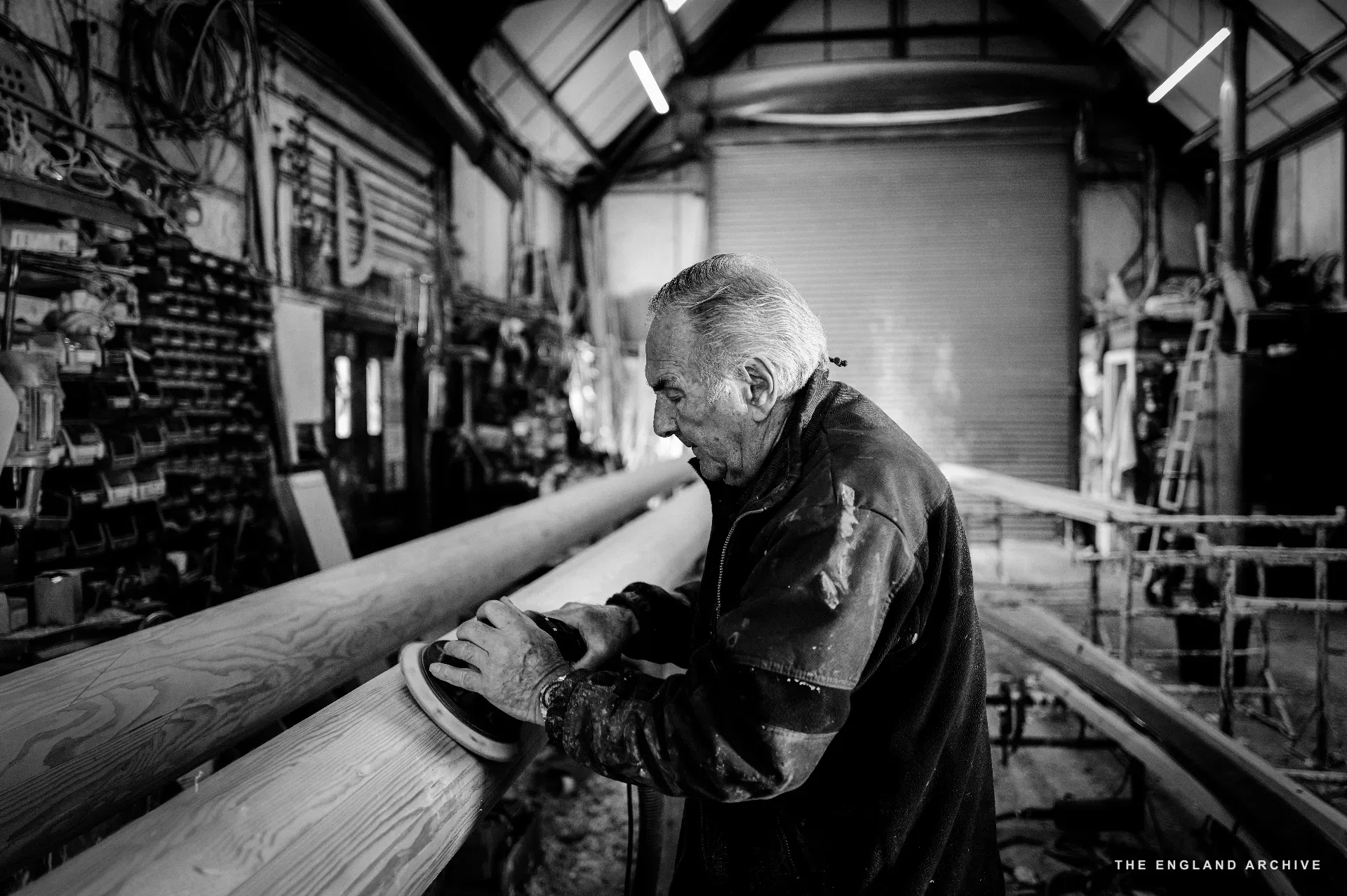 Michael Dennett working a long timber at the bench with an electric sander, the workshop’s side wall behind him with the door letting in flat outdoor light.