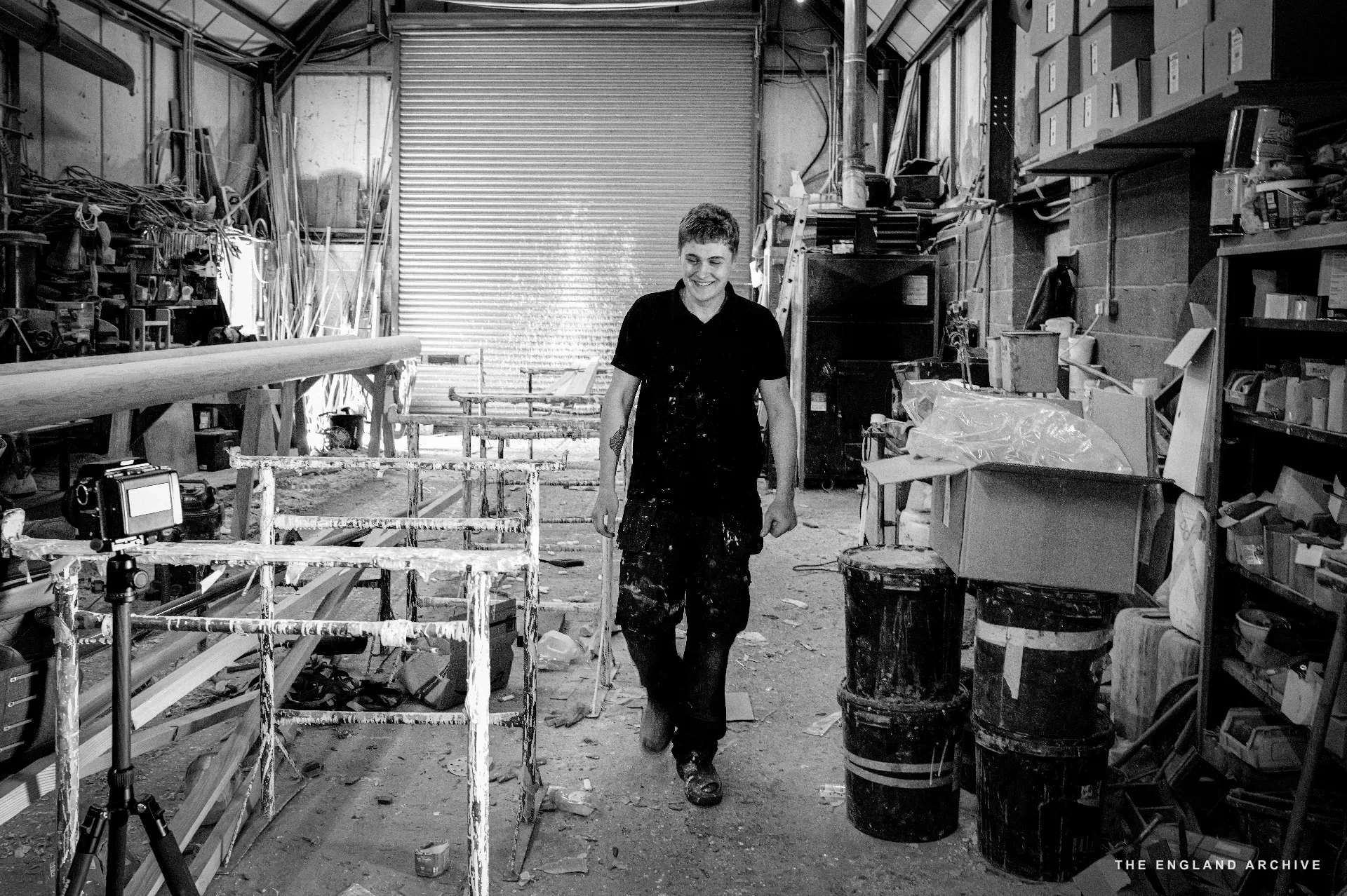 A worker walking down the central aisle of the workshop between rows of paint-encrusted boat trestles and the tool shelves, the high roof and corrugated wall behind.