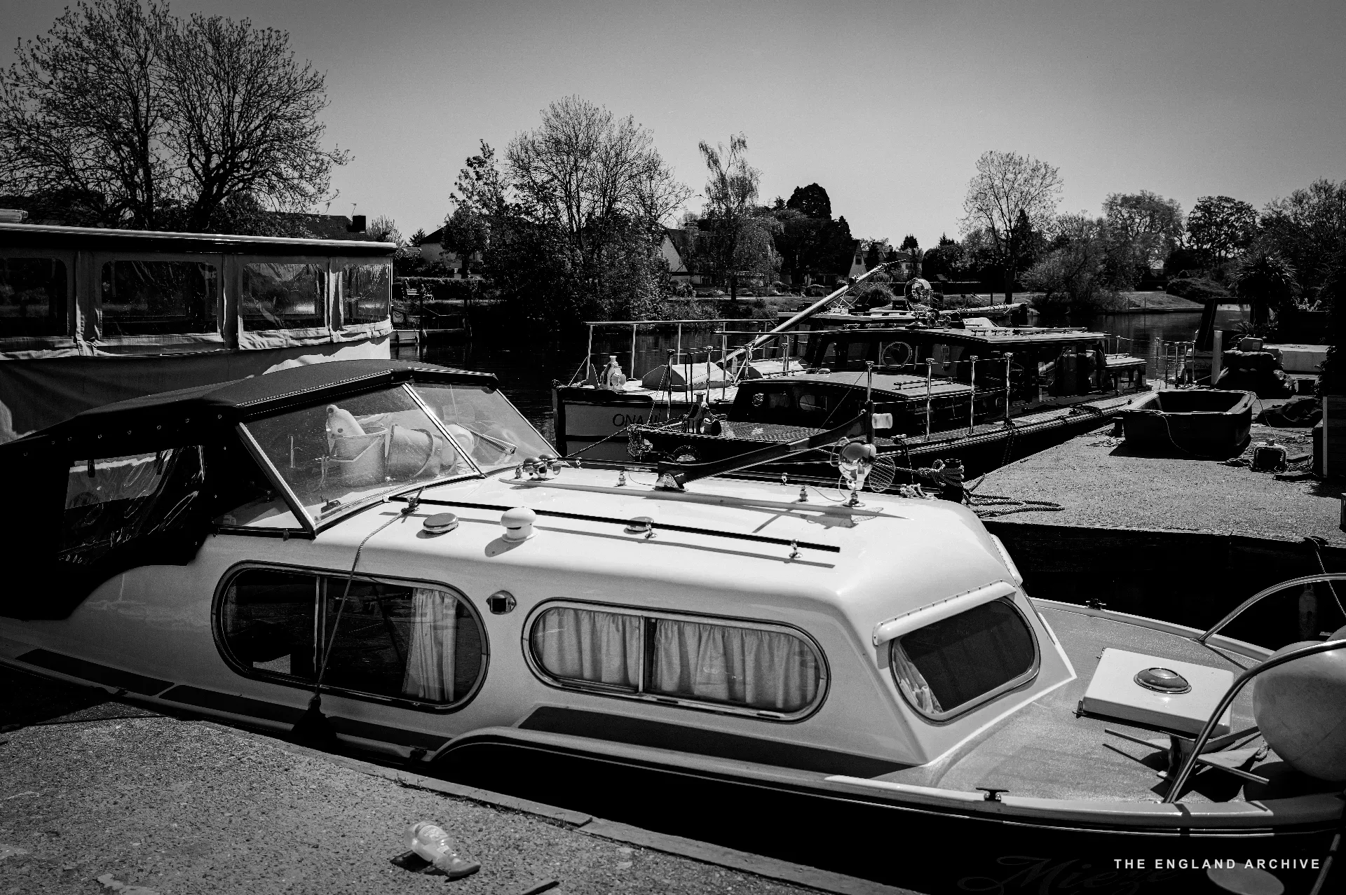 A line of moored boats at the yard’s slipway, the white river launch in the foreground with its cabin canopy raised, a sailing yacht and other vessels behind, willows and far houses across the river.