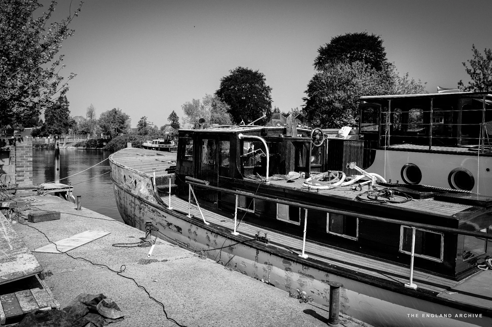 A long wooden river launch moored alongside the yard’s slipway, its cabin and brass detail catching the sun, the river curving past with willows and houses on the far bank.