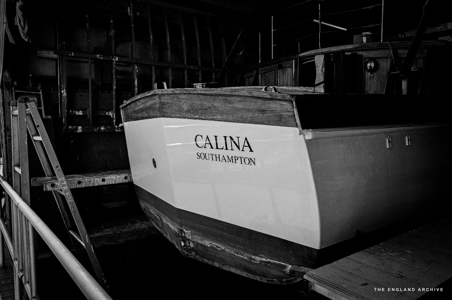 The bow of the boat ‘Calina, Southampton’ in the dark of the workshop, the painted nameplate clearly visible on the white hull. A wooden ladder propped against the cradle on the left.