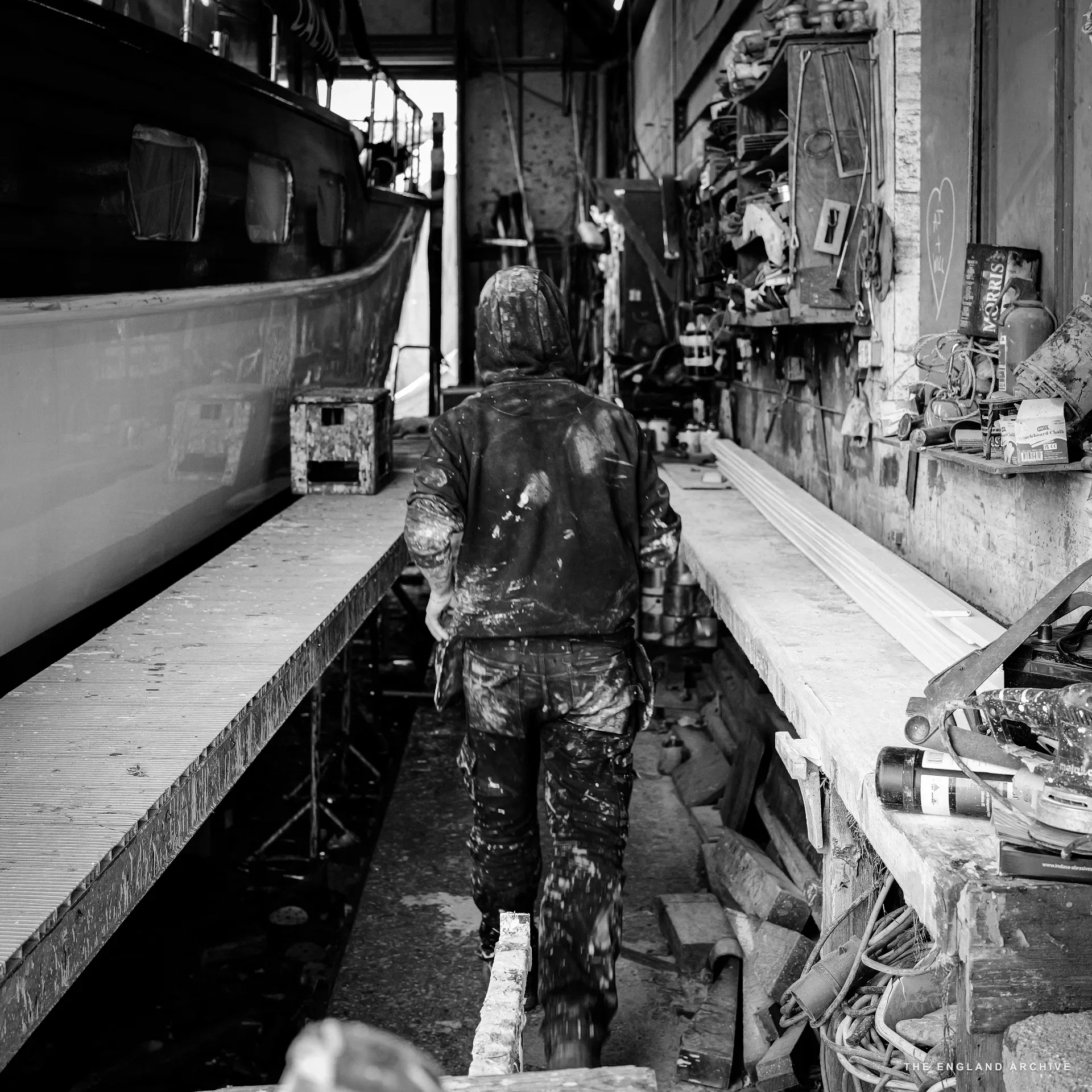 The young hooded apprentice walking down the long workshop aisle away from the camera, the side of a hull on the left, the bench and tool shelves on the right, the workshop receding to a bright doorway at the far end.