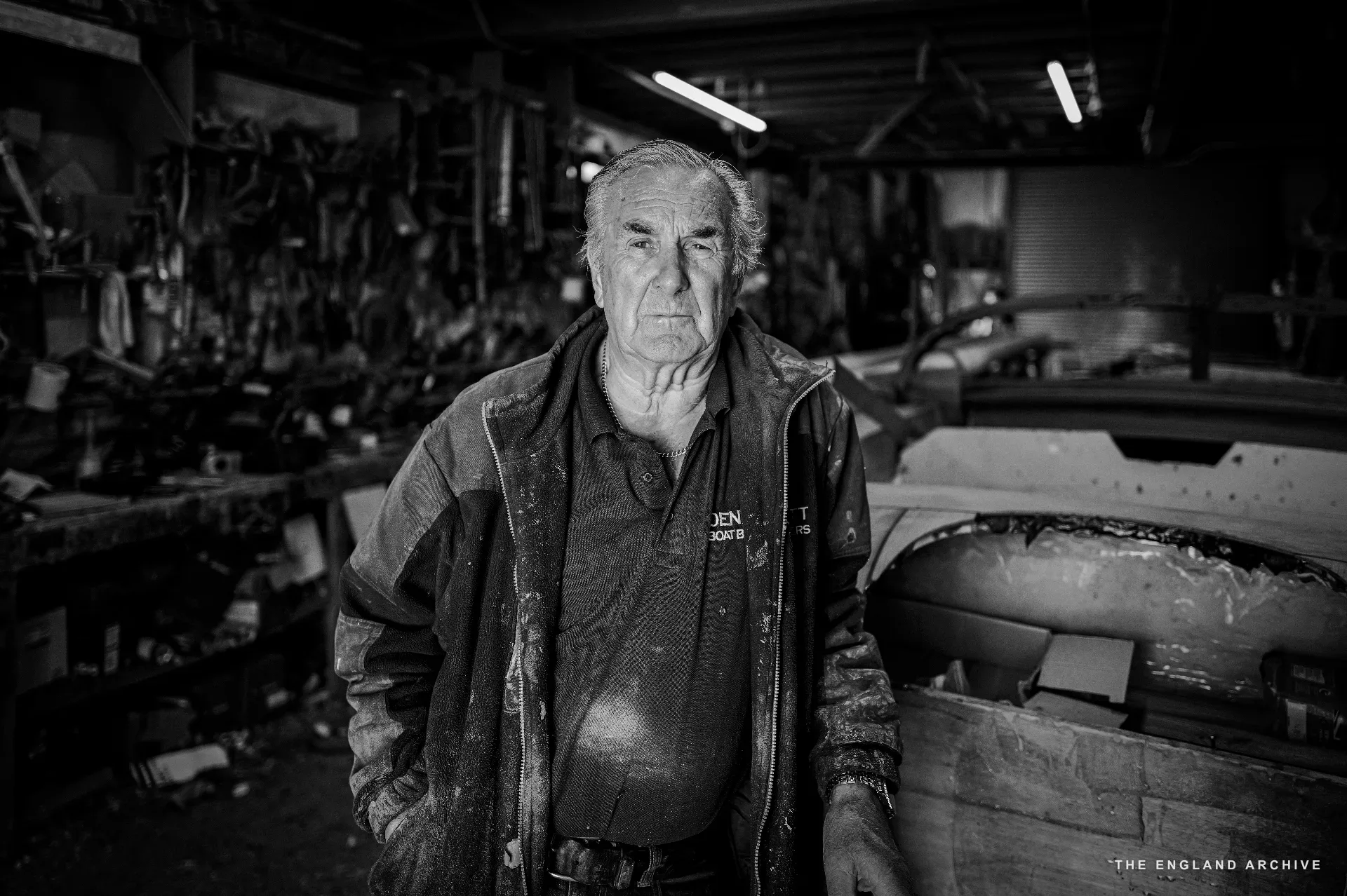 Michael Dennett standing in the workshop in his work jacket, looking at the camera, hands in his pockets, the tool rack and a partially built hull visible behind him.