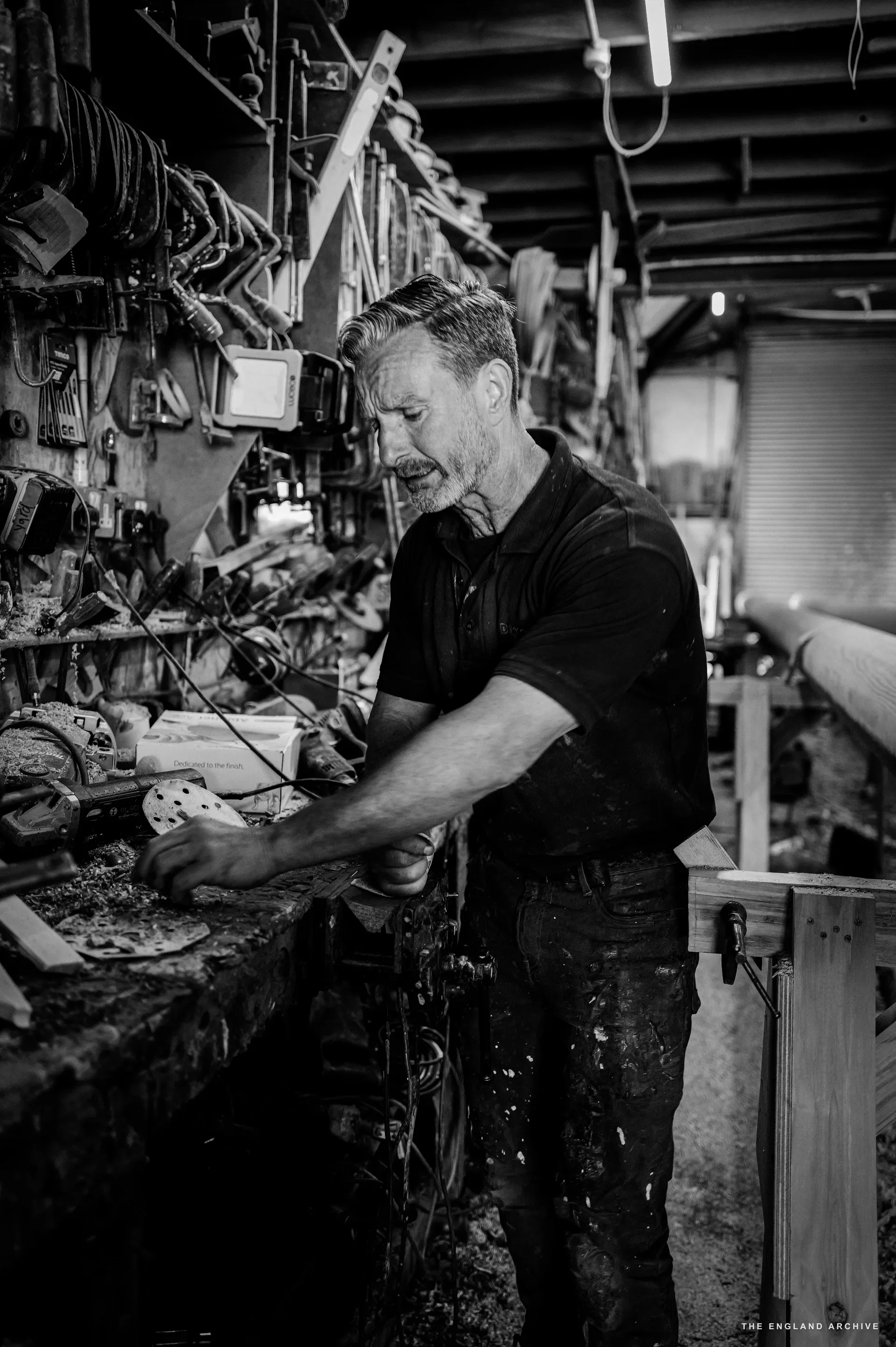 Stephen Dennett at the bench, side profile, the dense tool wall and shelves behind him, working with a small tool in his right hand.