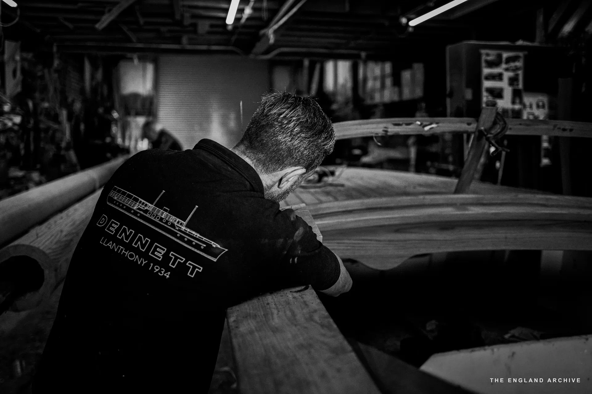 Stephen Dennett (back to camera, the DENNETT LLANTHONY 1934 polo across his shoulders) bent over a stack of long timbers in the workshop, planning the next cut.