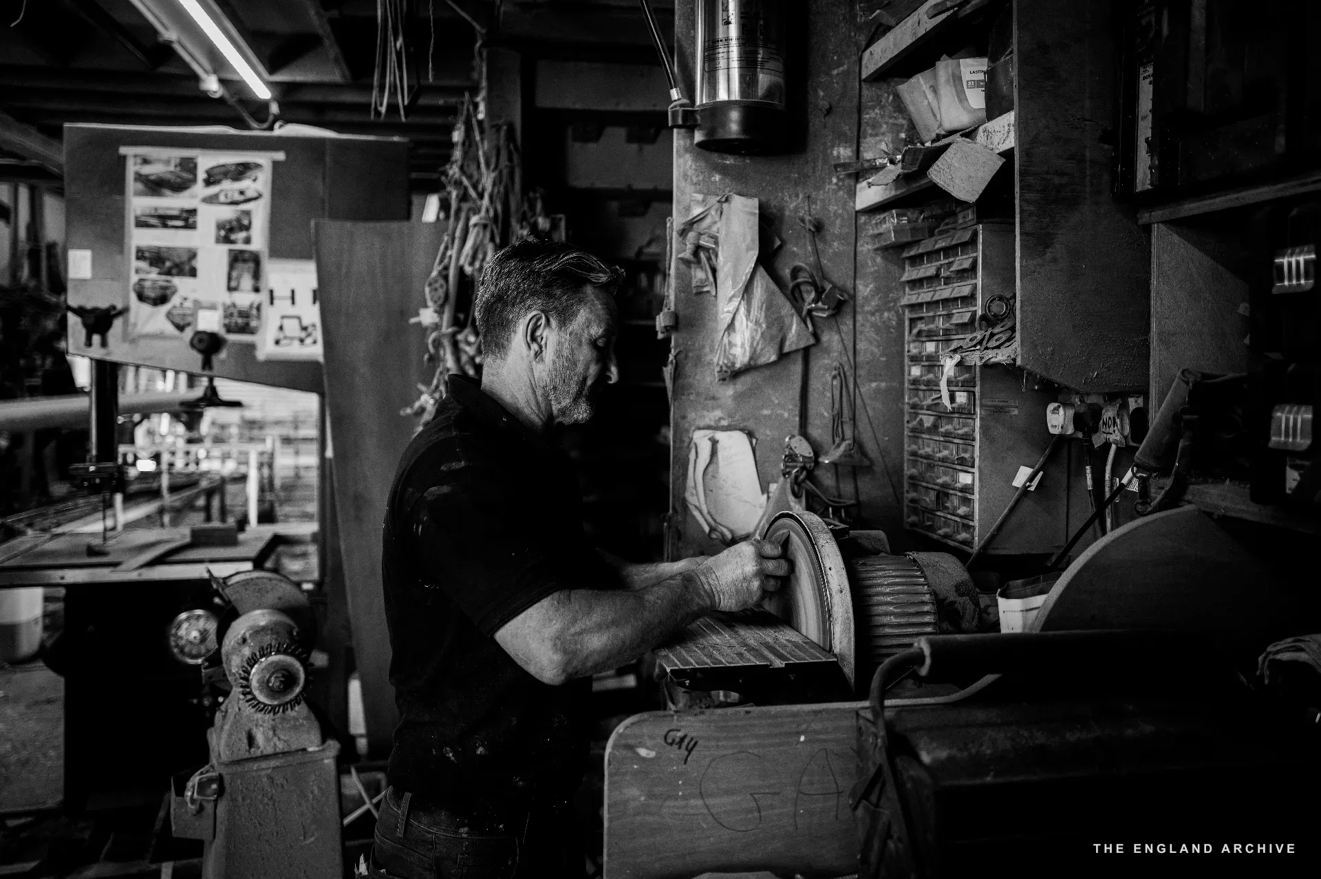 Stephen Dennett at the bench using a circular grinding wheel on a small piece of timber, the tool wall and a vintage compressor visible behind him, fluorescent strip lighting overhead.