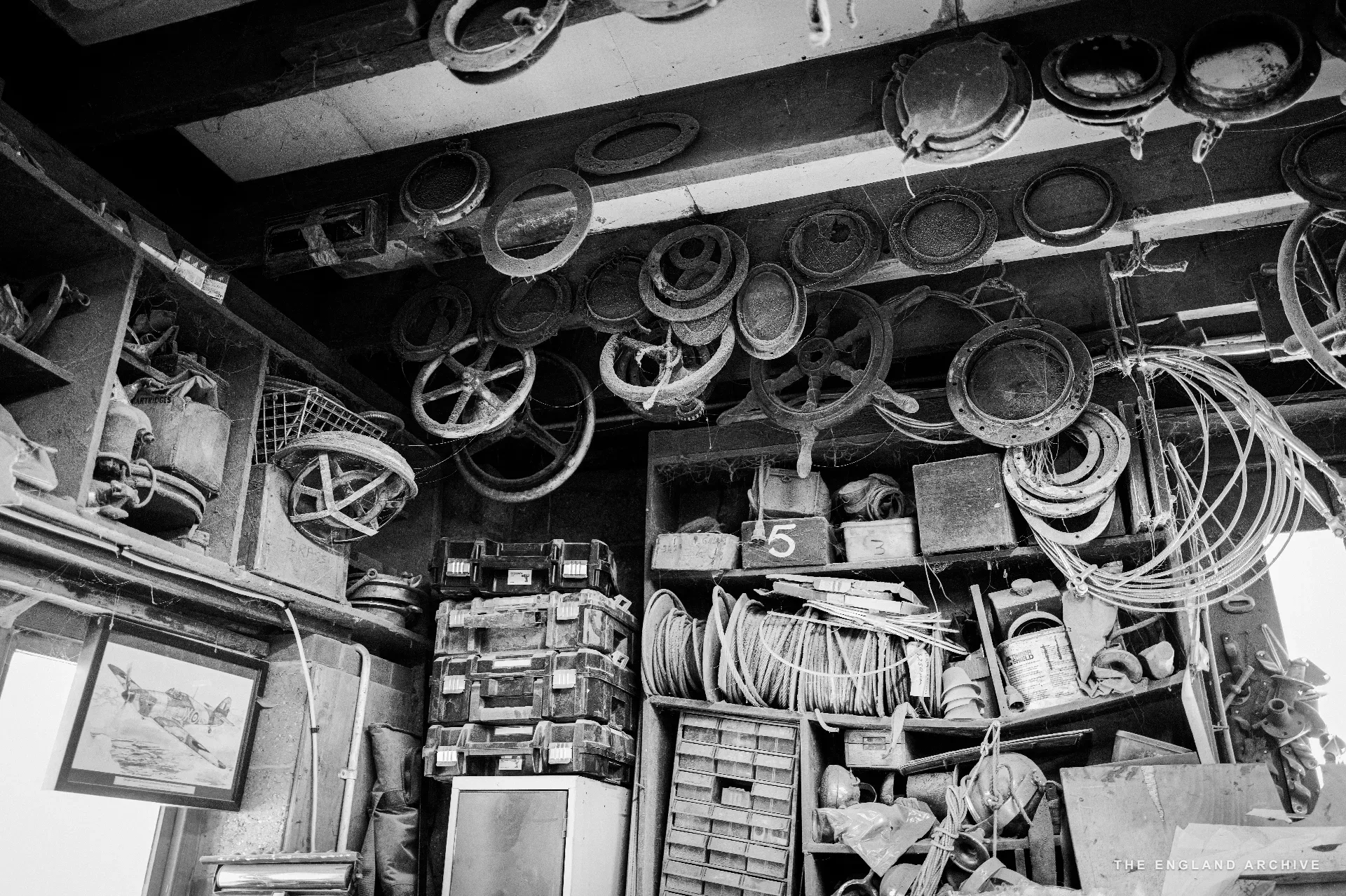 A workshop corner: dozens of cast-iron porthole frames hanging from the rafters above a bench piled with lengths of cable, brass fittings and small drawers - the parts archive of a yard that has rebuilt every kind of river boat.