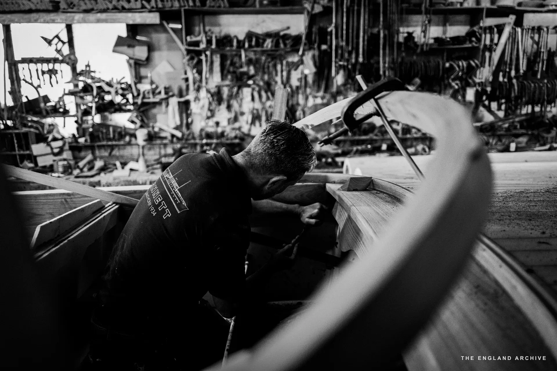 Stephen Dennett standing inside the open hull of a small boat under construction in the workshop, working a curved timber on the rim, the tool shelves visible behind.