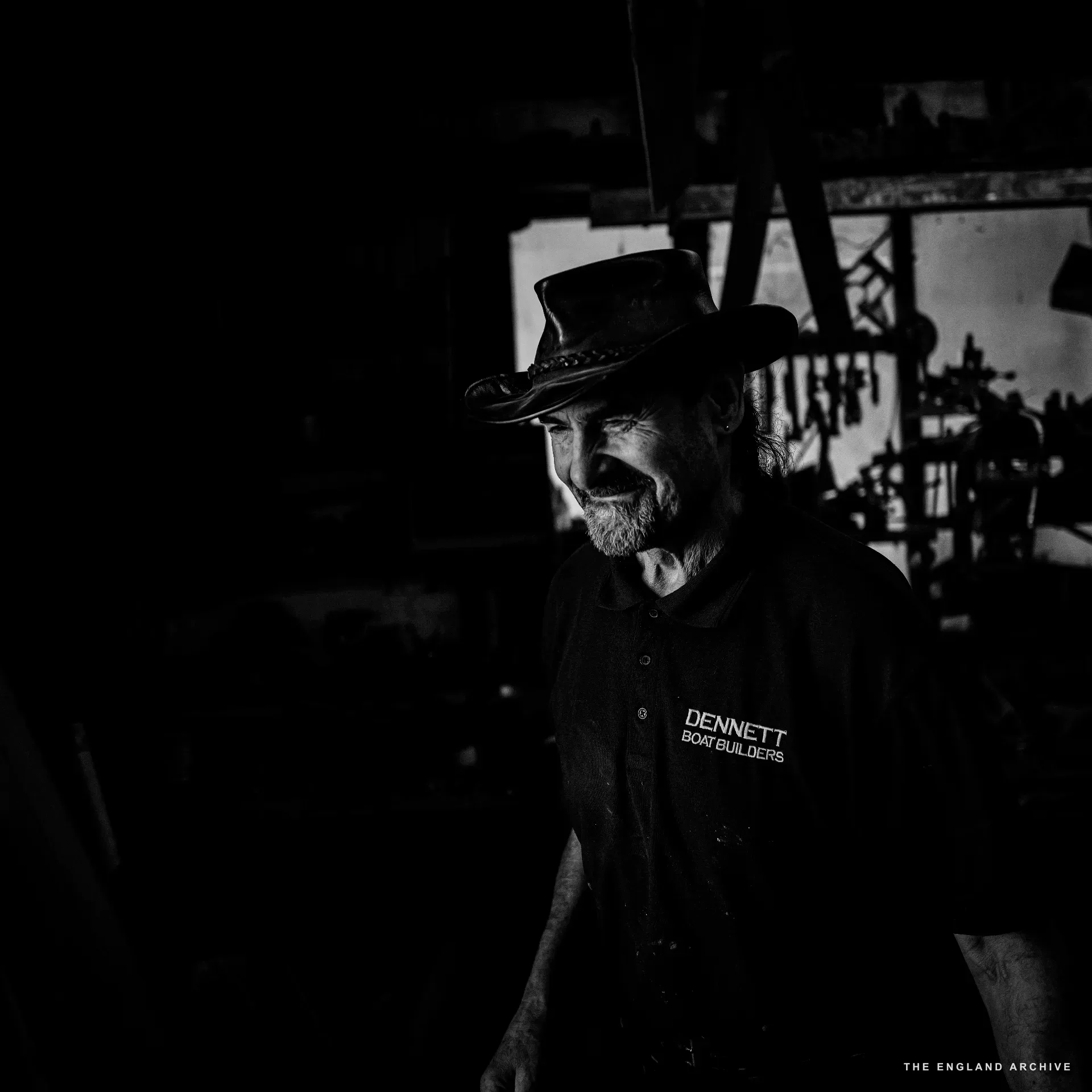 A low-key portrait of the leather-hatted yard worker in the workshop - DENNETT BOAT BUILDERS polo just visible, soft daylight from the right, the workshop dark behind him.