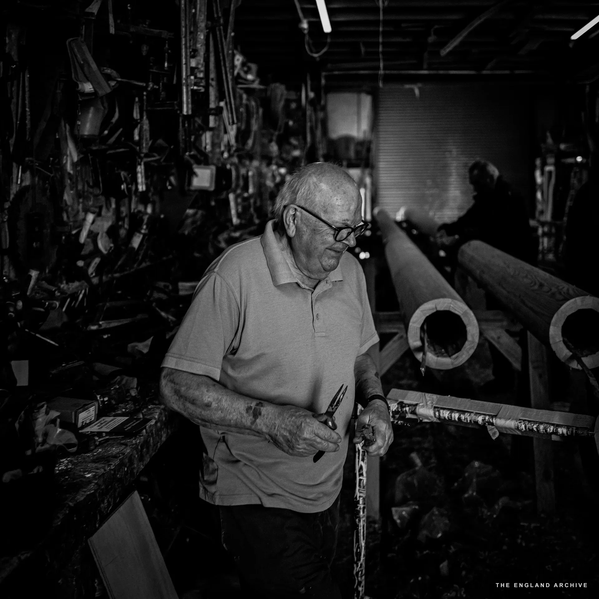 An older worker in glasses standing at the workshop’s tool wall, working a small piece in his hands, a row of long timbers laid on trestles behind him.