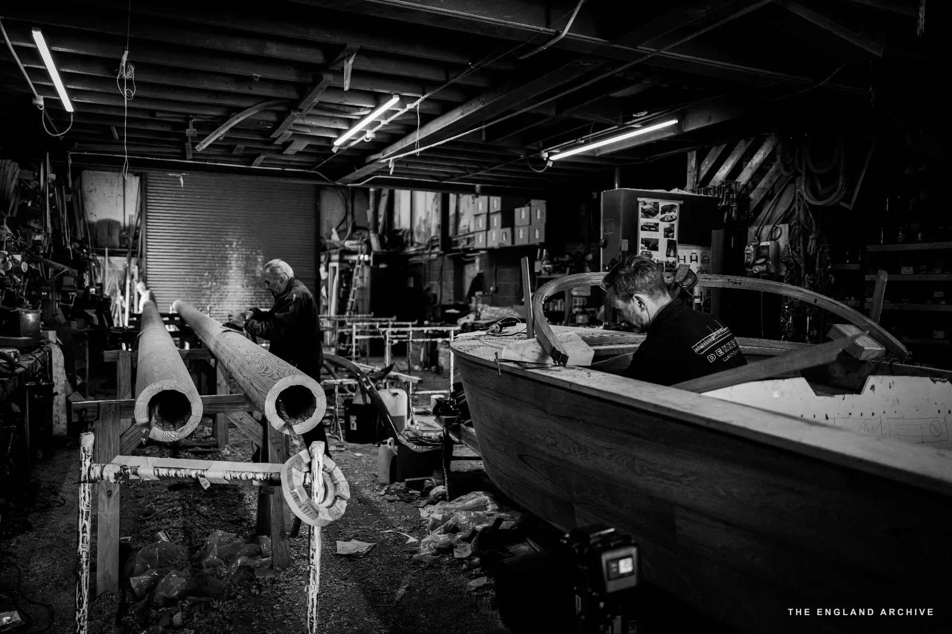 A wide view of the workshop with two workers visible - one standing at a long timber on a trestle in the middle distance, another bent over the deck of a small boat on the right. The corrugated workshop walls rise behind.
