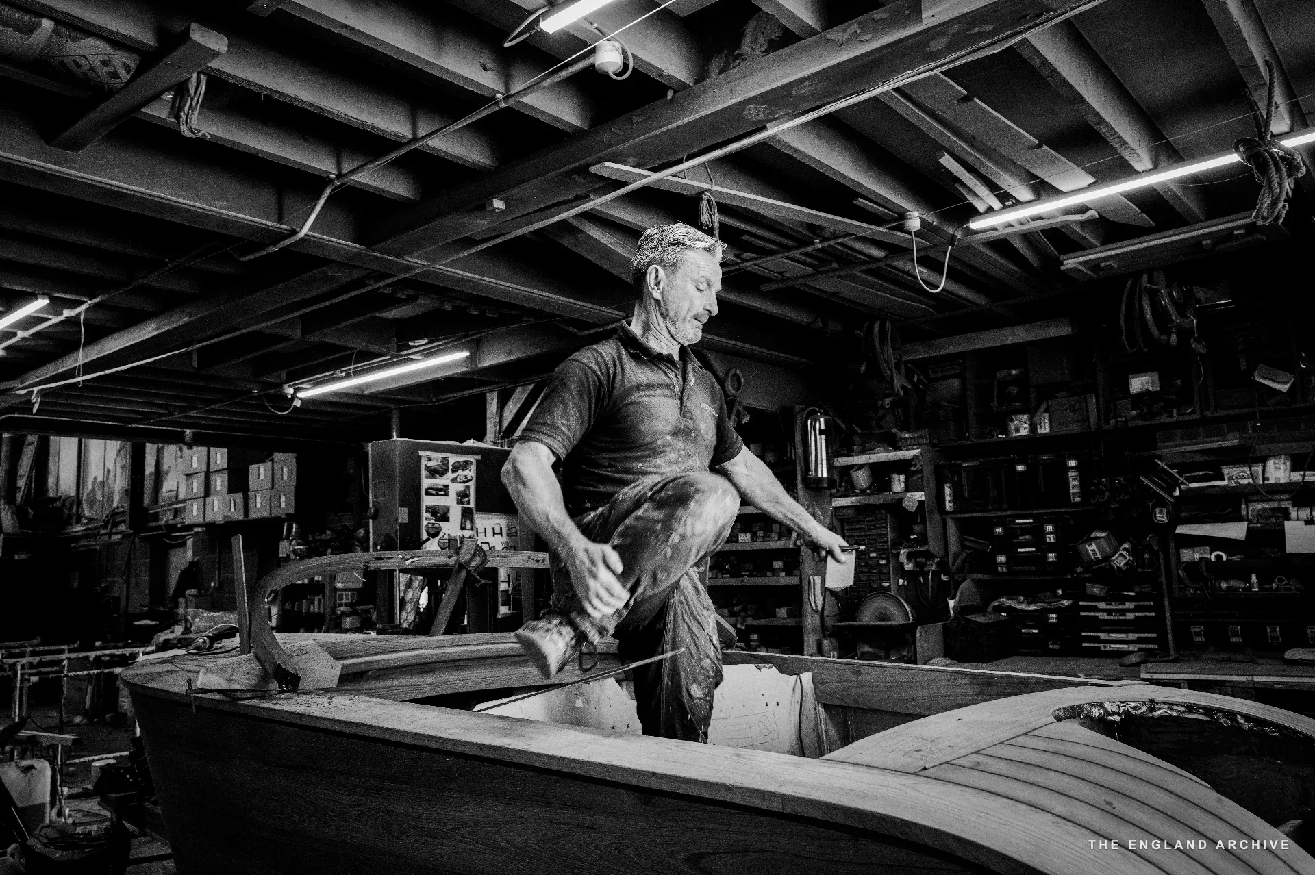 Stephen Dennett stepping over the gunwale of a small boat in the workshop, mid-stride, hands raised for balance, the bench and tool shelves filling the background.