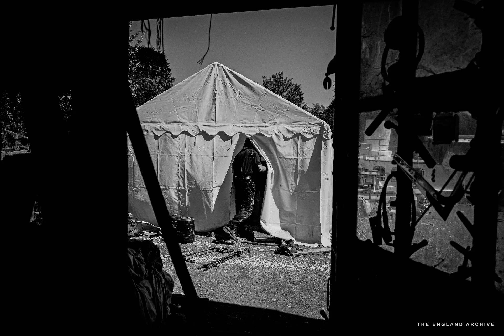 A high-key view through a workshop doorway out into the yard - a white canopy tent set up in the sunlight, a worker just visible inside the tent, the dark interior of the workshop framing the door.