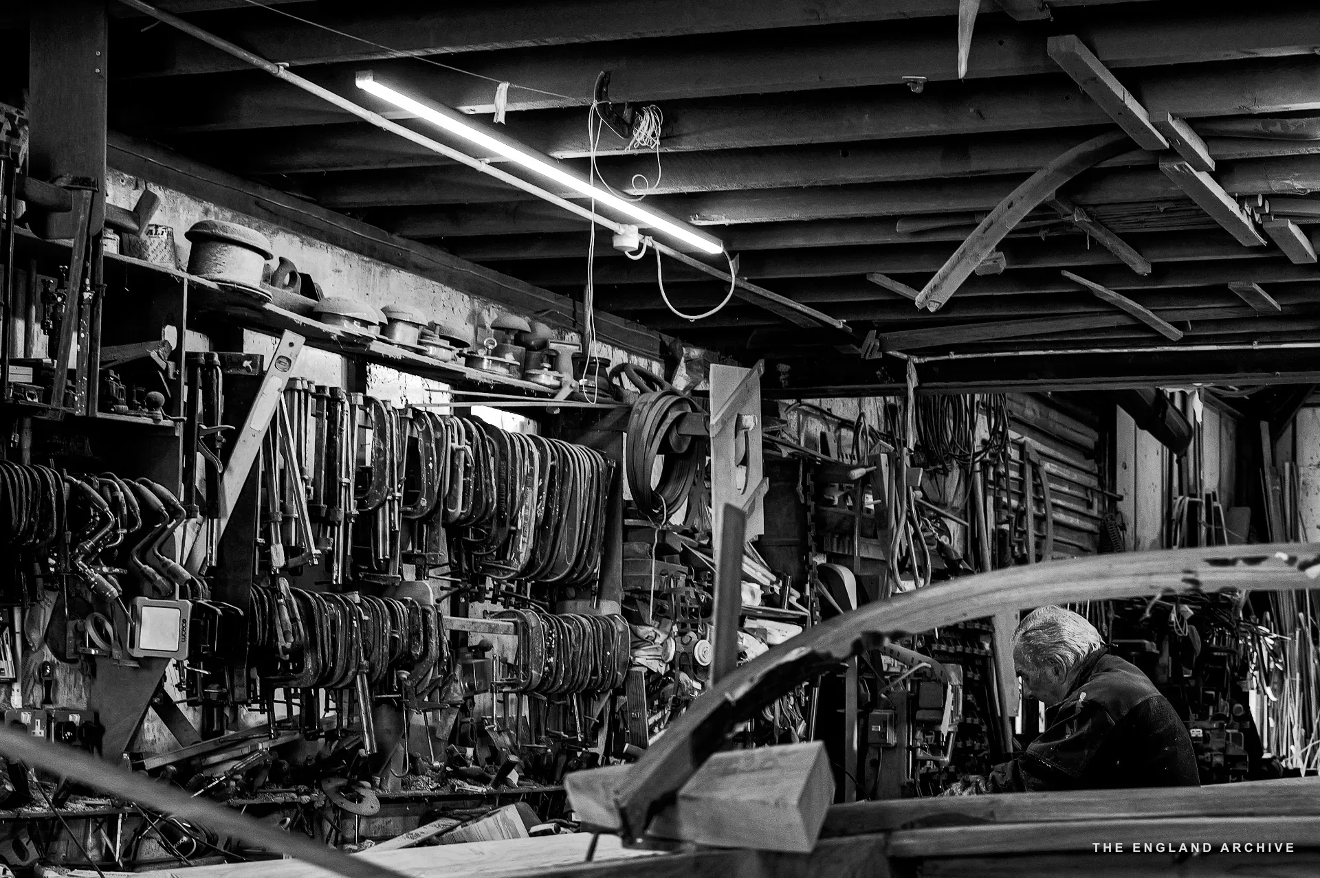 A wide view of the workshop’s side wall - a fluorescent strip light over the bench, dense tool racks, a hull on the right edge of frame, and a worker dimly visible at the far bench.