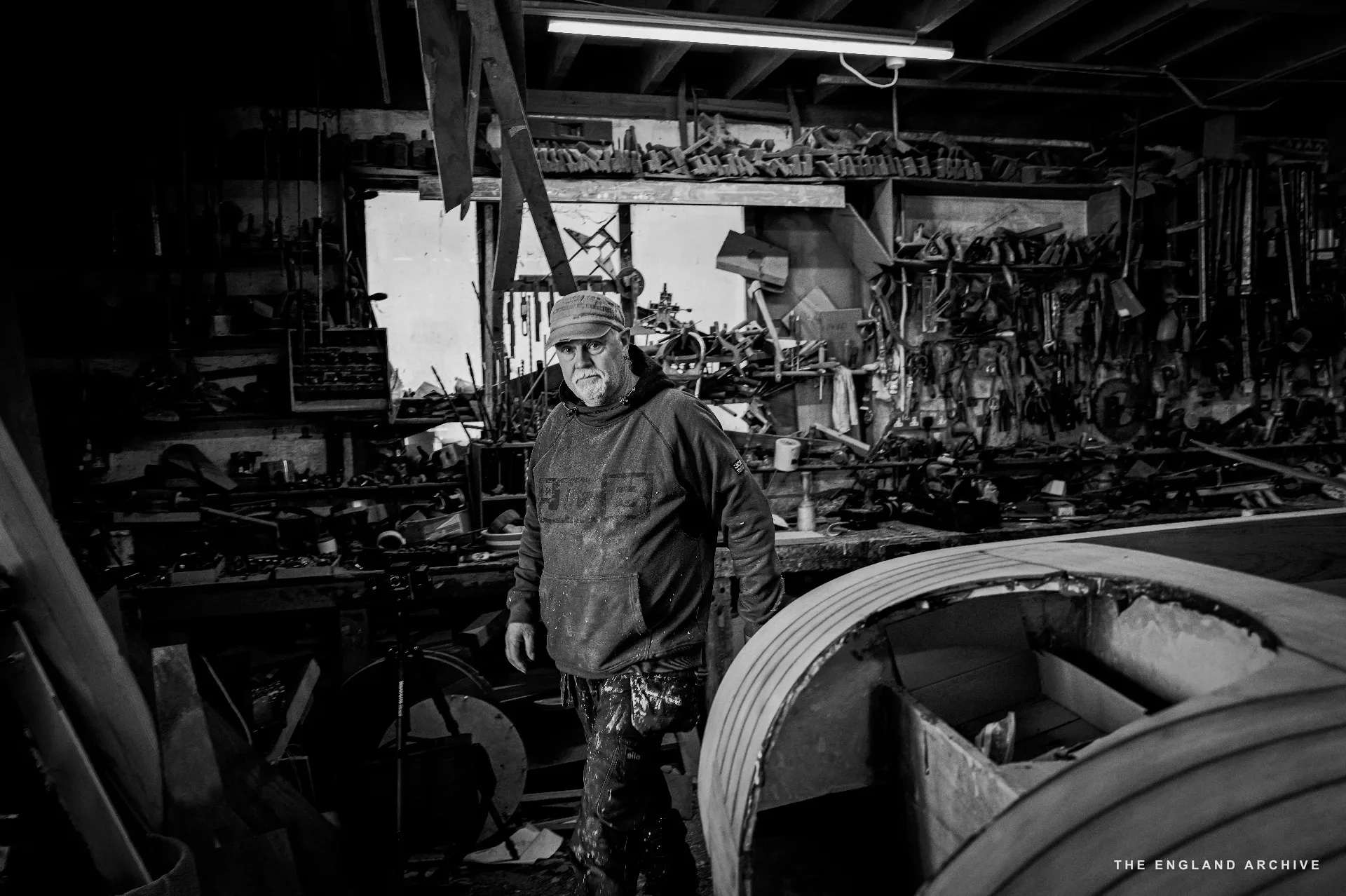 A worker in cap and dark jacket standing in the workshop next to a partially planked hull, looking past the camera, the tool wall stretching to either side.