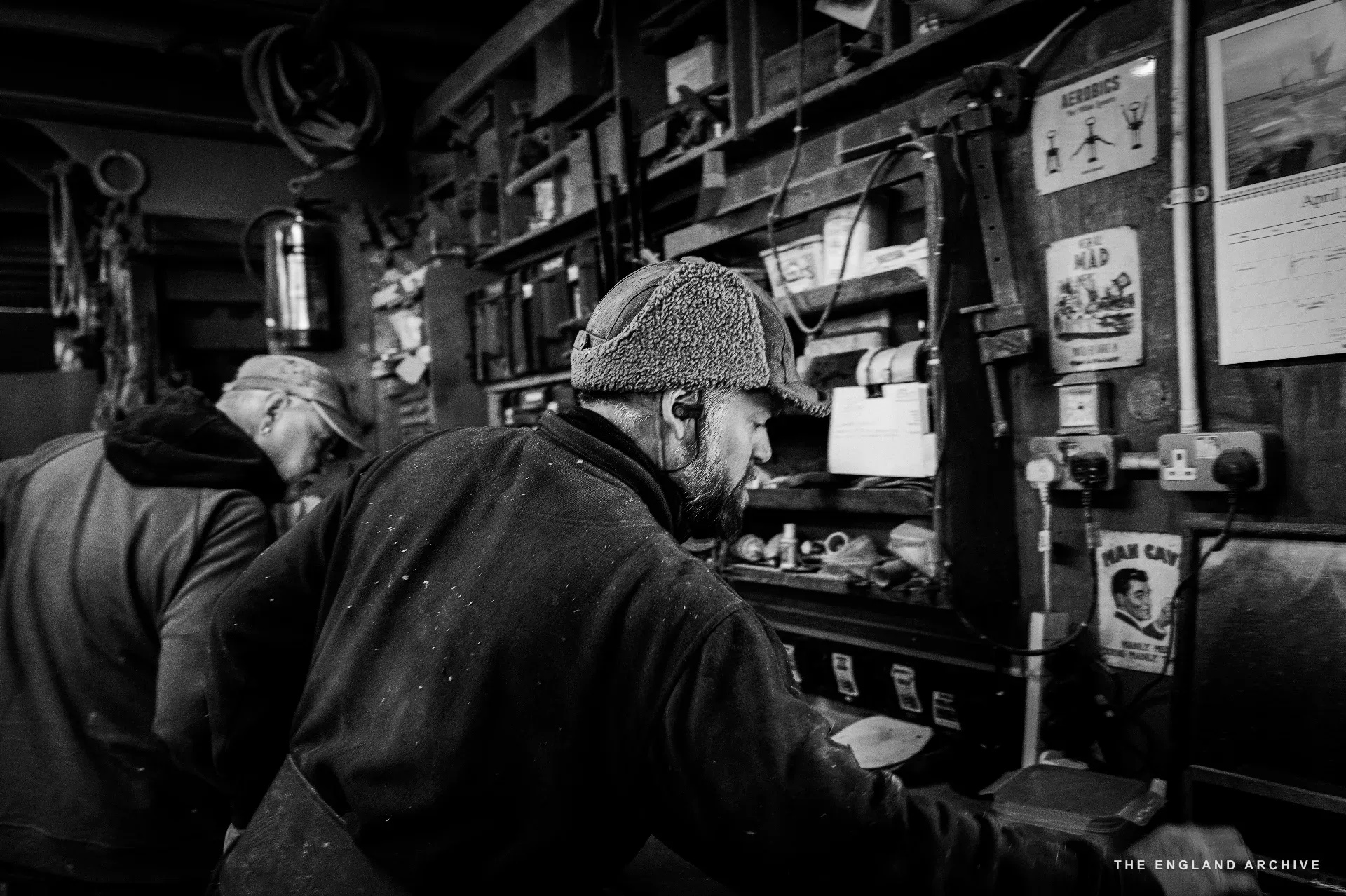 The leather-hatted yard worker (centre) at the workshop bench, head turned slightly, two other workers behind him at separate tasks, walls of pinned posters and tools to the right.