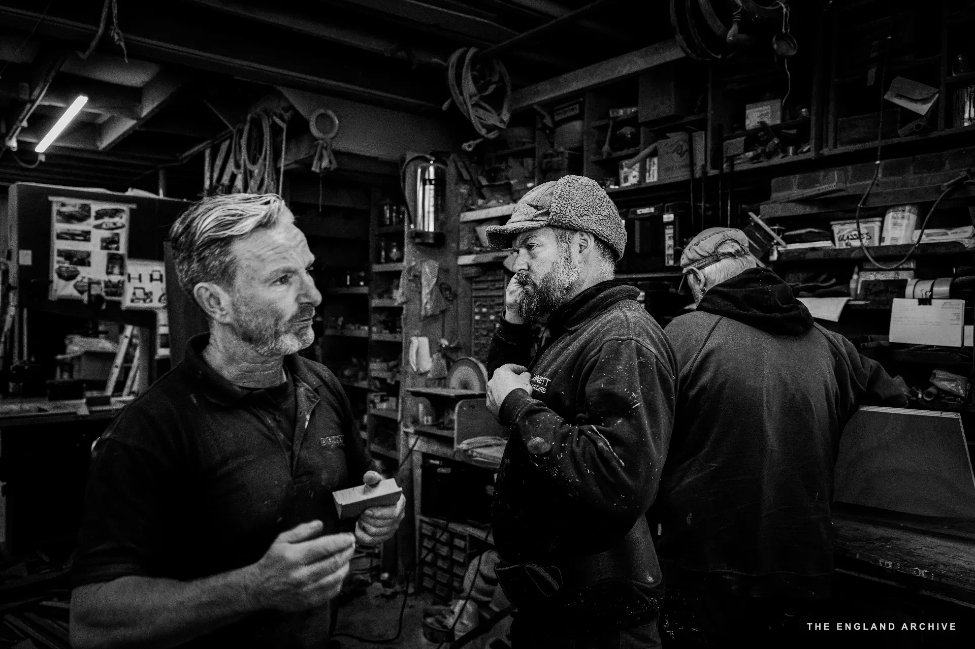 Stephen Dennett (left, in DENNETT polo) in conversation with the leather-hatted yard worker (centre) and a third worker (right, back to camera), the tool wall and shelves rising behind them.