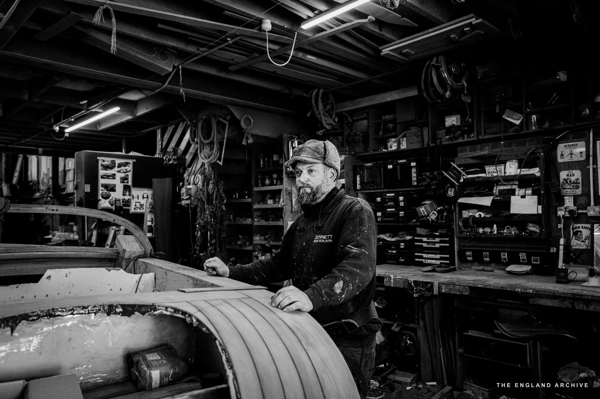 A bearded worker in cap and DENNETT polo standing inside the open hull of a boat under construction, the planking curving up around him.