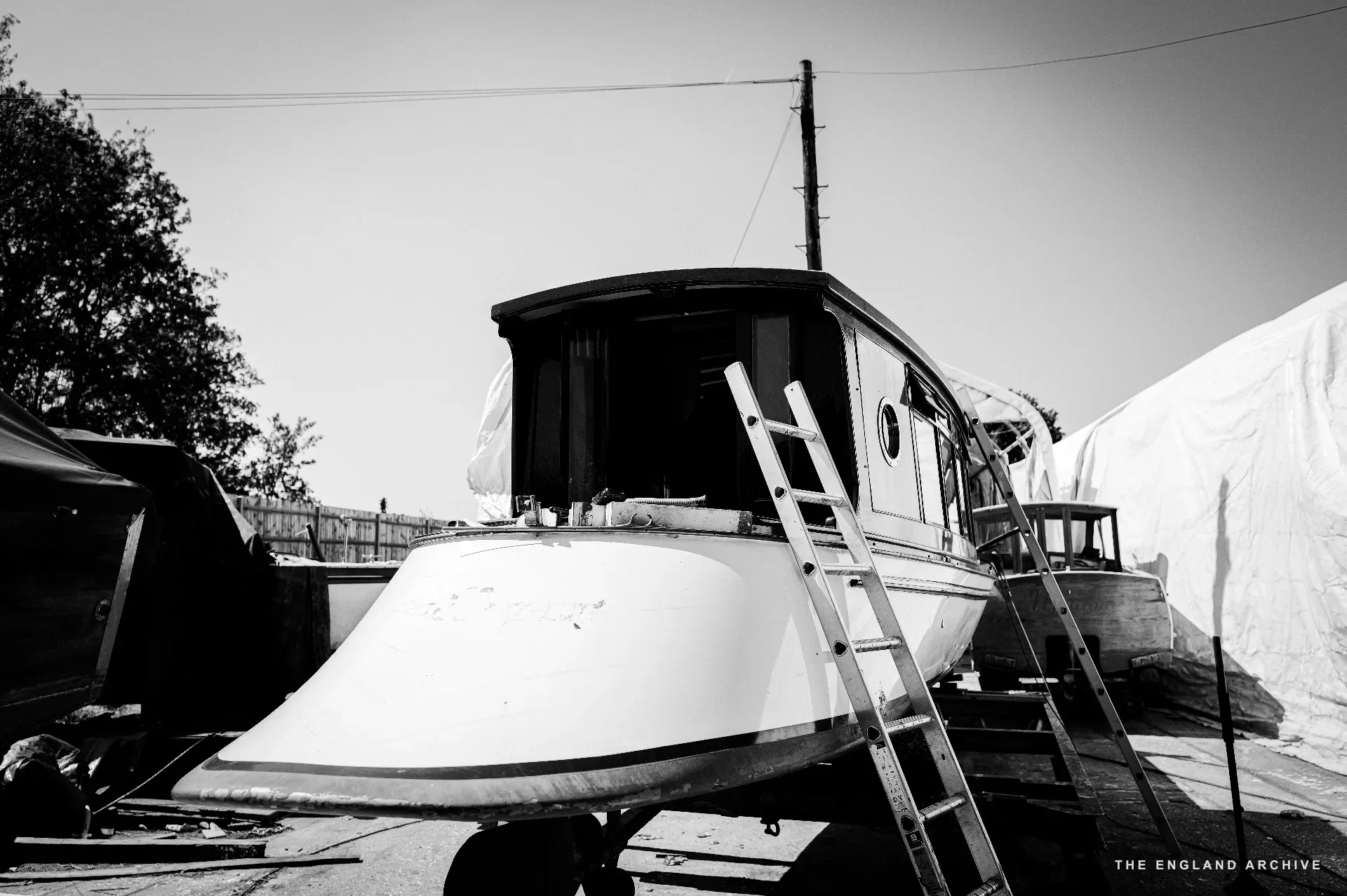 A tall wooden cabin cruiser propped on chocks in the yard, a wooden ladder leaning against its side, a tarpaulin tent behind it.