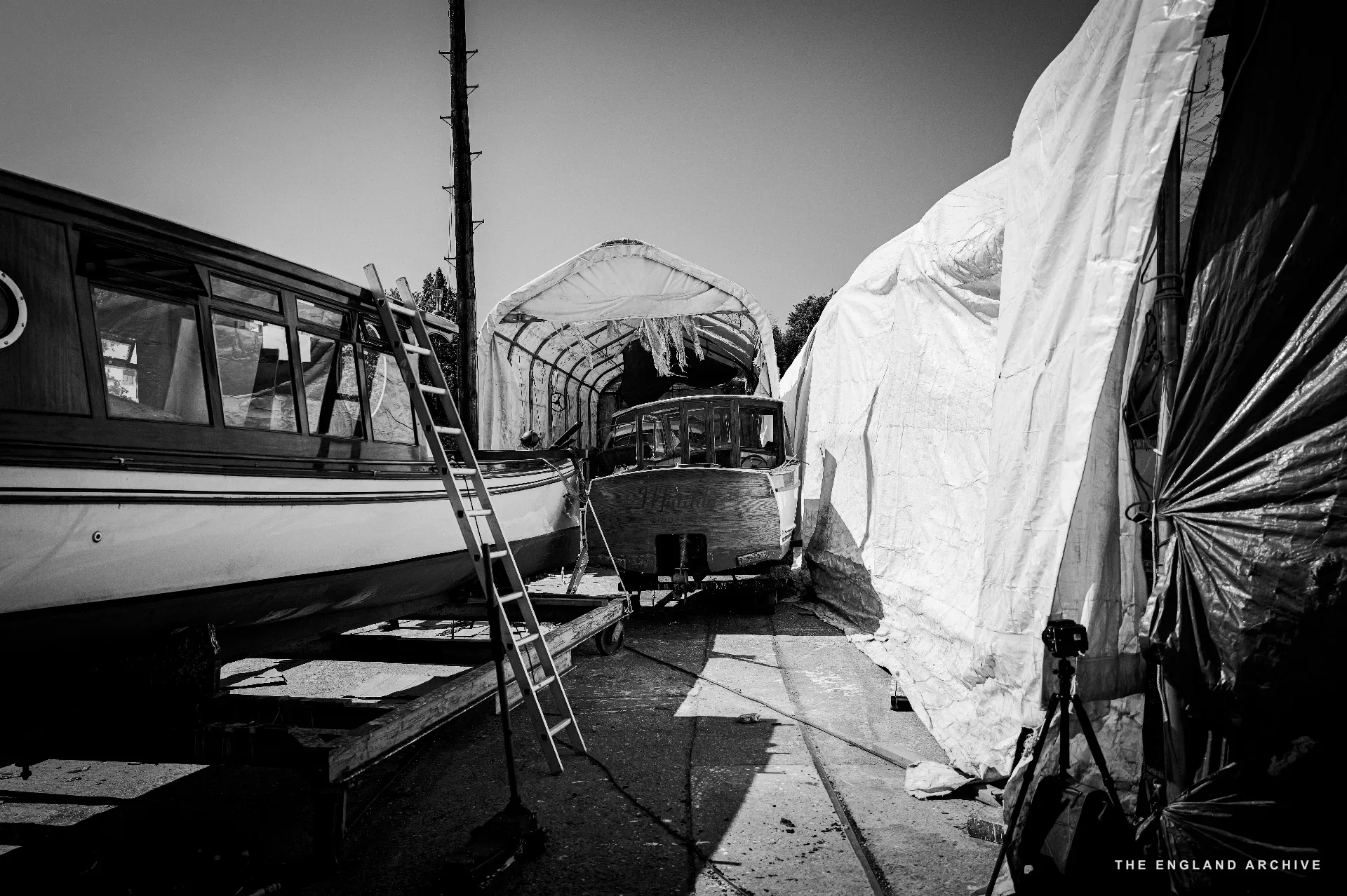 A wide yard scene - several boats on chocks under tarpaulin tents, a ladder and a tripod in the foreground, the riverside trees at the back.