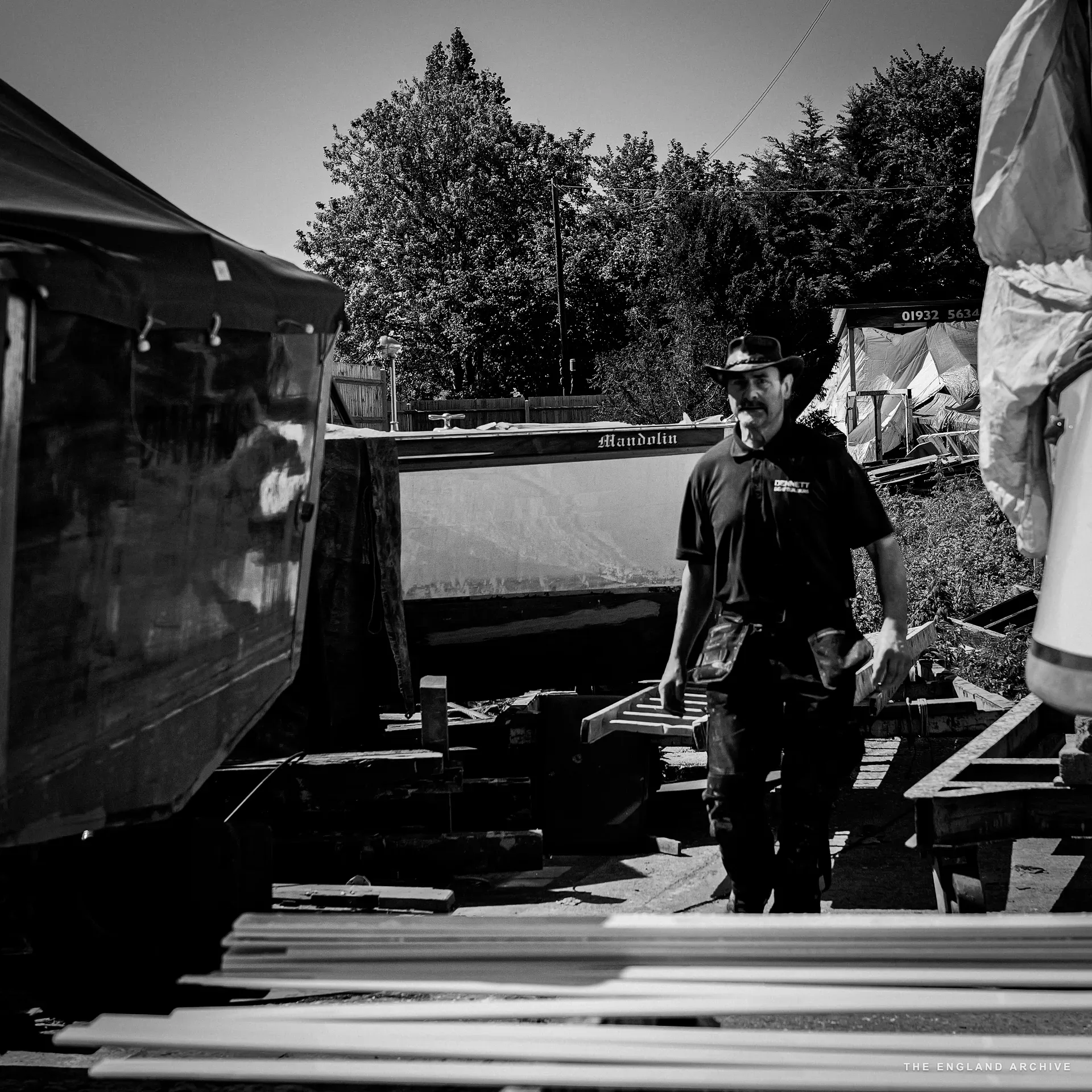 The leather-hatted yard worker walking through the yard, the boat ‘Mandolin’ on chocks behind him, the workshop building visible to the right.