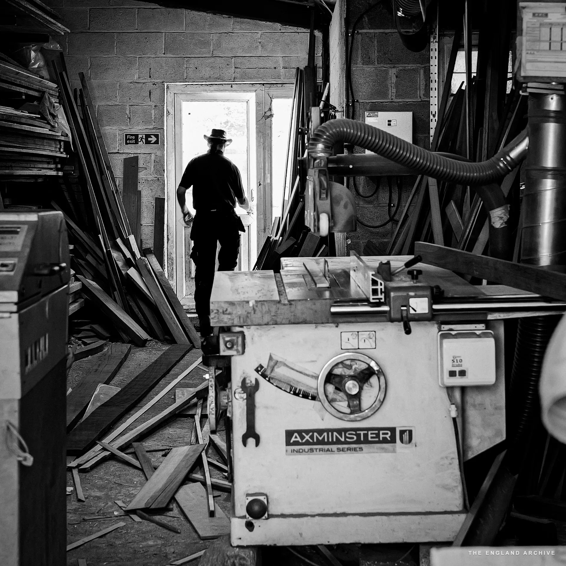 The leather-hatted yard worker walking out of the workshop into a bright doorway, the Axminster industrial saw bench in the foreground and rows of tall timber boards leaning against the workshop walls.