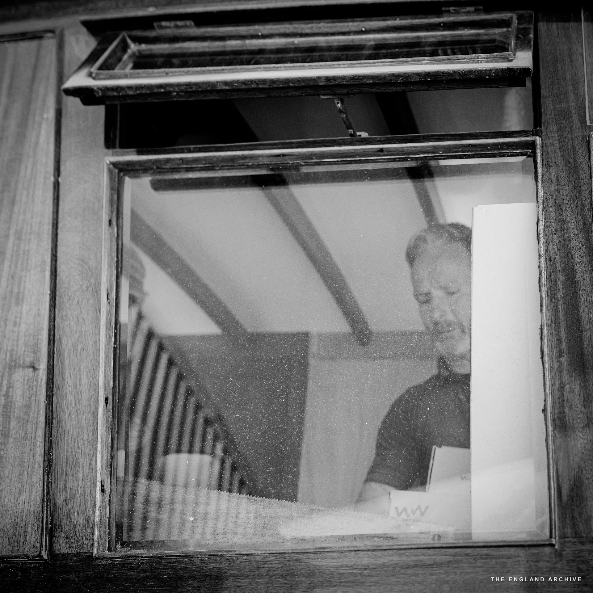 A worker (Stephen Dennett) seen through the cabin window of a boat in the workshop, head down on paperwork at a small table inside the cabin, the wooden ceiling beams of the cabin behind him.