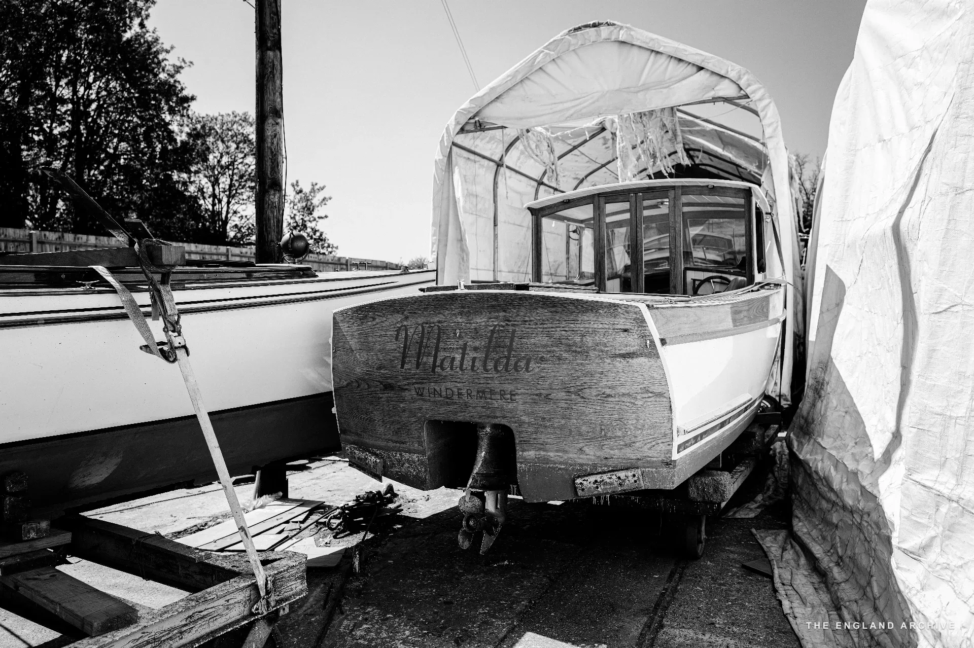 The boat ‘Matilda’ on a trailer in the yard under a tarpaulin tent, name and lettering visible on the stern, the workshop building behind.