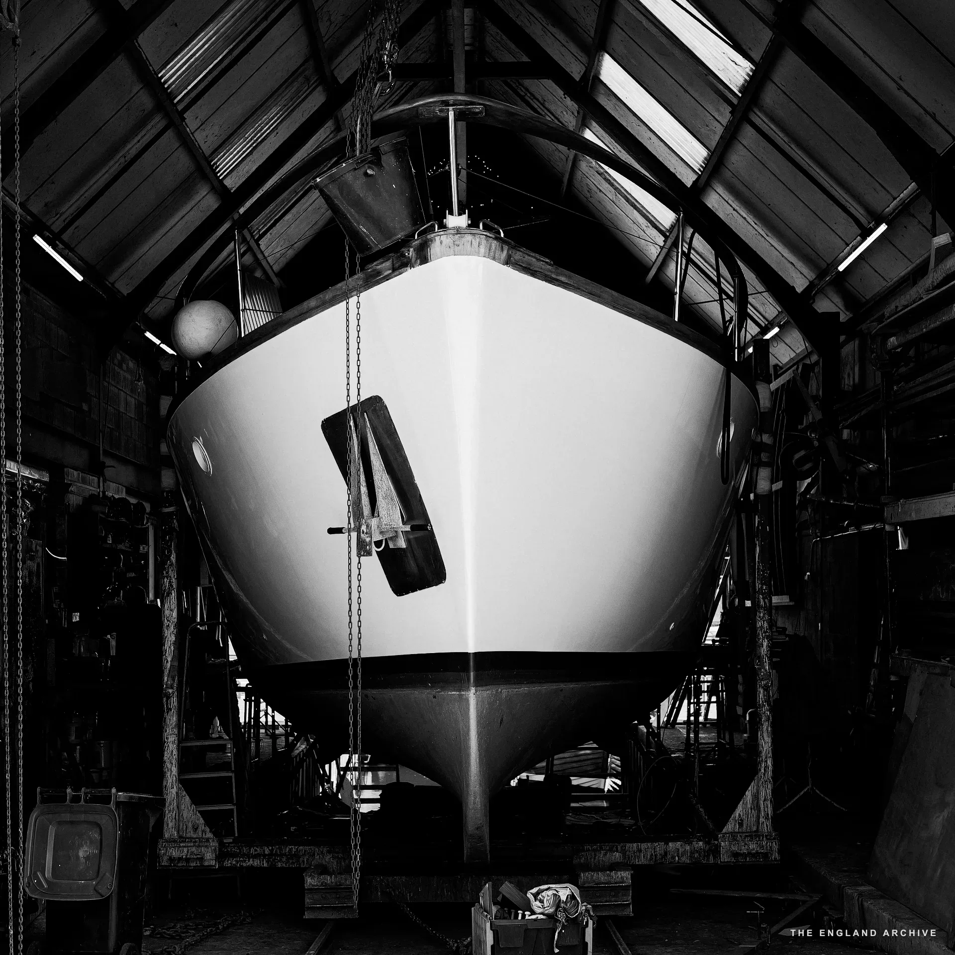 A sailing yacht under construction in the workshop’s tall main shed, viewed bow-on, the rudder hanging on chains in the centre, the boat’s white hull catching the daylight from the high pitched roof above.