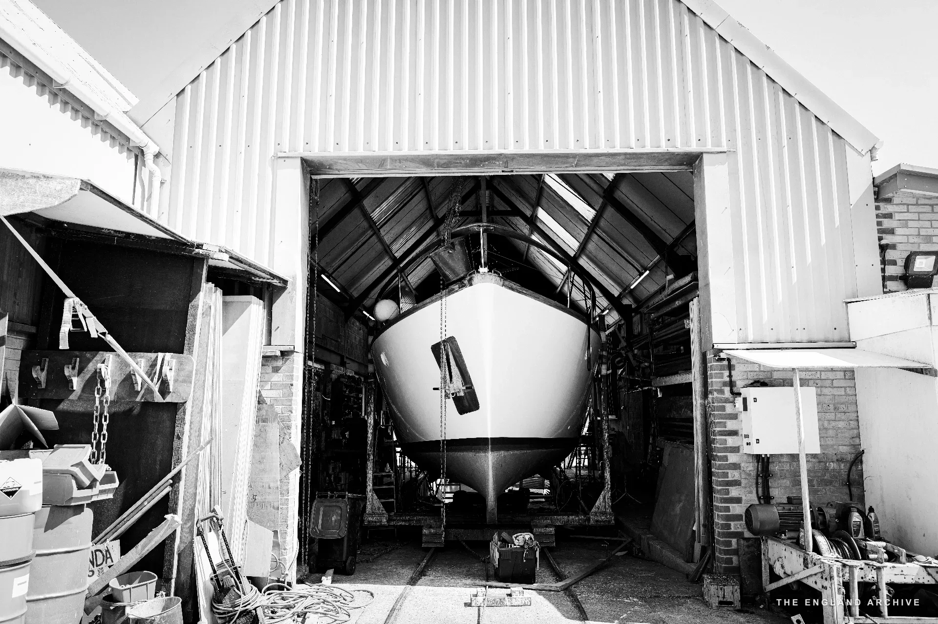 The same sailing yacht in the workshop seen from the doorway - the boat propped up in the centre of the shed with the open doors framing it, rusted wheelbarrows and equipment to either side.
