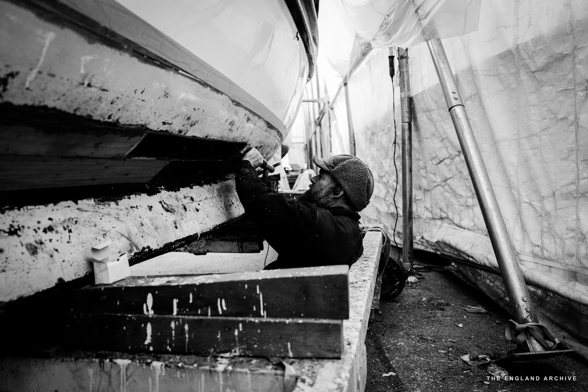 A worker in a knitted cap working under the side of a hull, hands raised on the planking, a tarpaulin tent and the workshop wall behind.