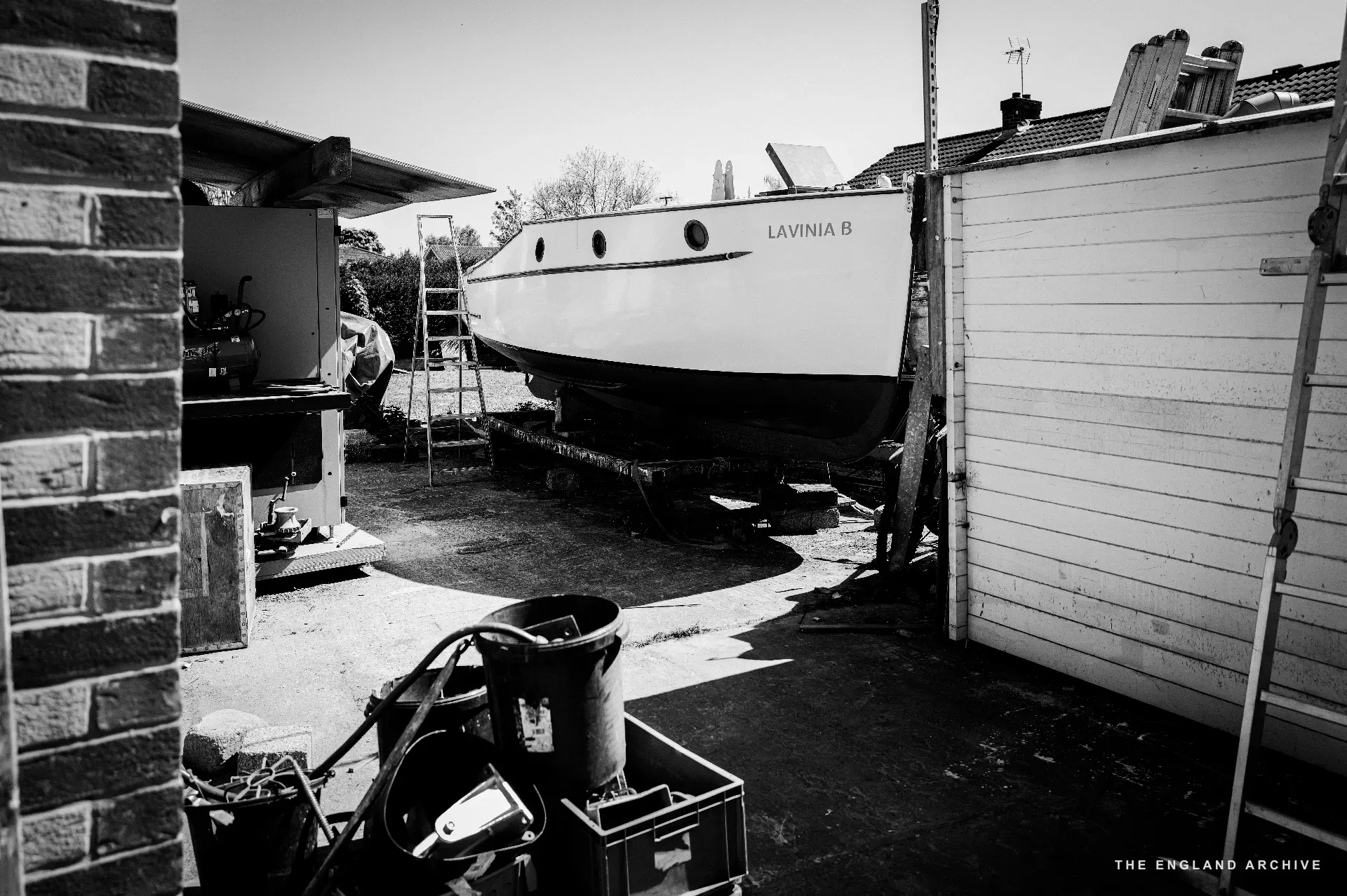 The boat ‘Lavinia II’ on a trailer in a narrow side yard between the workshop building and a neighbouring house, a wooden ladder leaning against the cabin and a metal water-tank in the foreground.