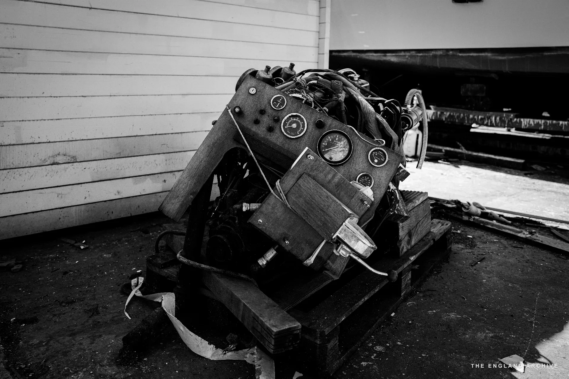 A vintage marine engine on a wooden pallet outside the workshop, polished brass instrument panel on its side, a corrugated workshop wall behind.