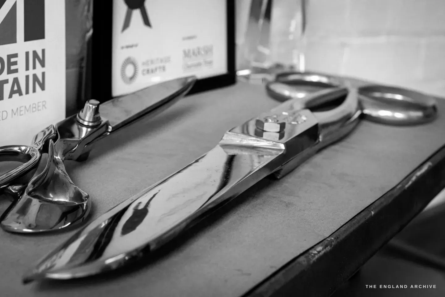 A pair of large polished tailor’s shears laid on a bench, with framed Made in Britain and Heritage Crafts member certificates and a glass award standing behind.