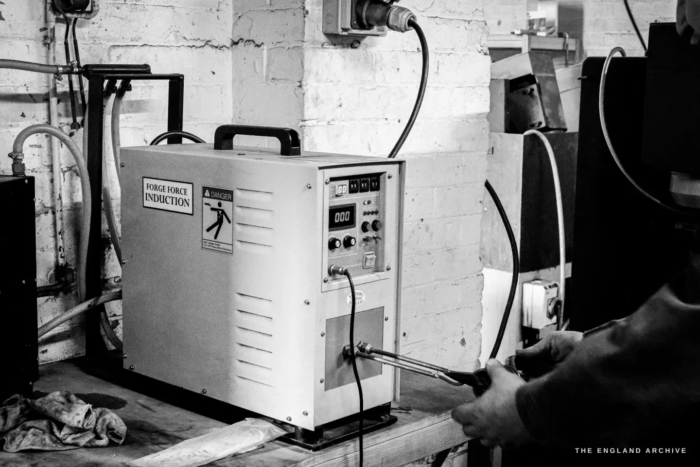 An induction annealing machine labelled FORGE FORCE INDUCTION against a whitewashed brick wall, a hand feeding a blade into the heating coil.