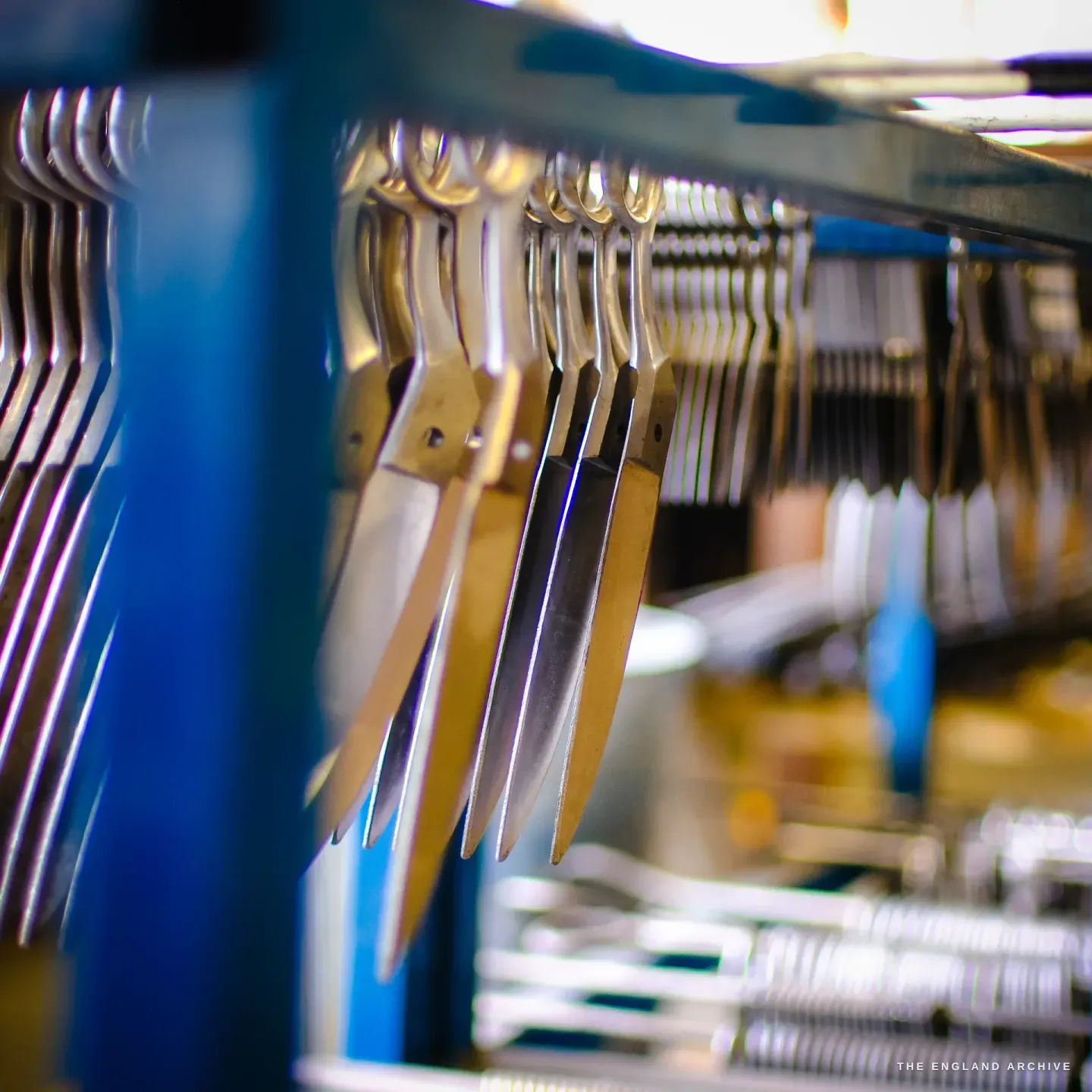 Rows of part-finished scissor blades hanging blade-down from a rack, the blue machine frame behind.