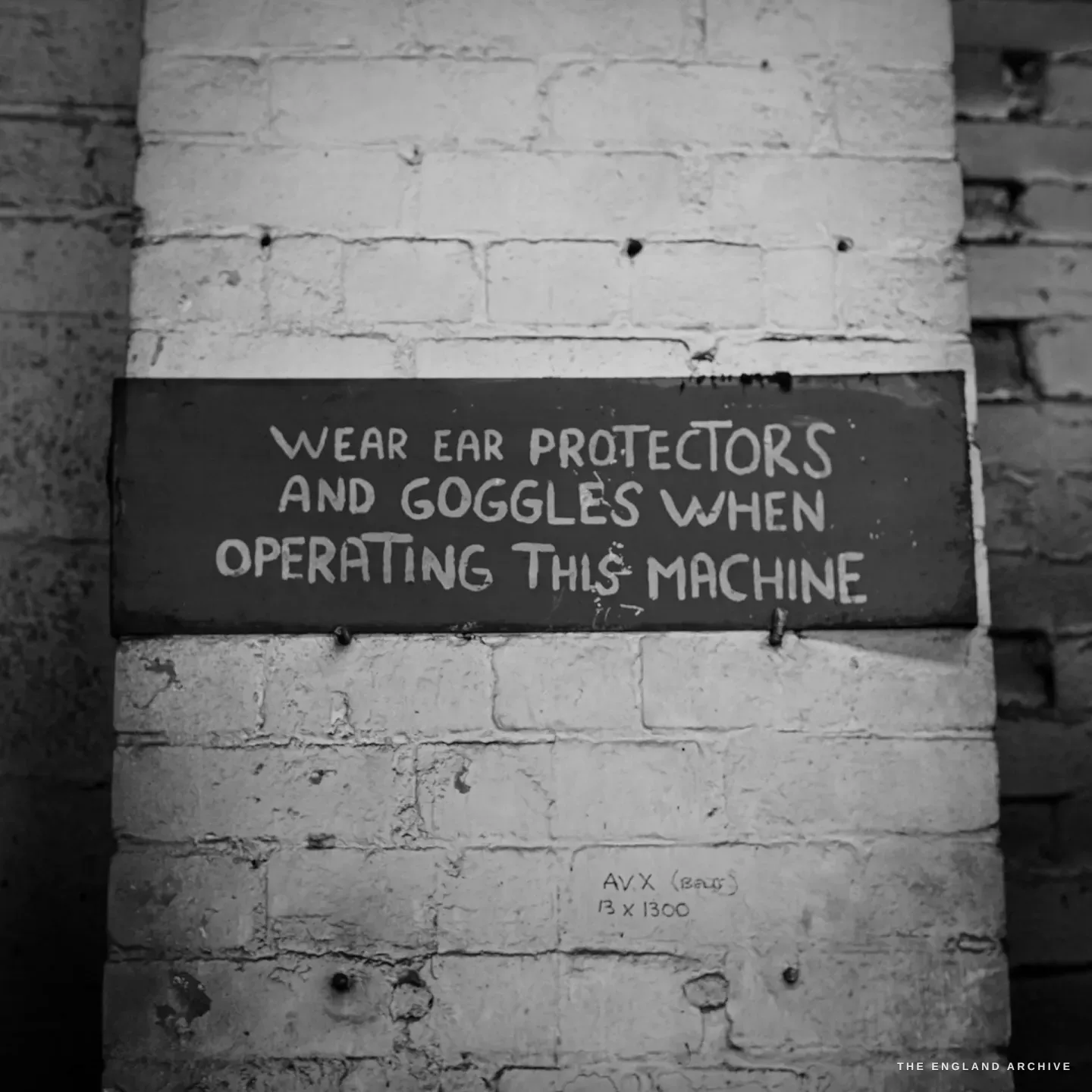 A hand-painted sign on a whitewashed brick pillar reading WEAR EAR PROTECTORS AND GOGGLES WHEN OPERATING THIS MACHINE.