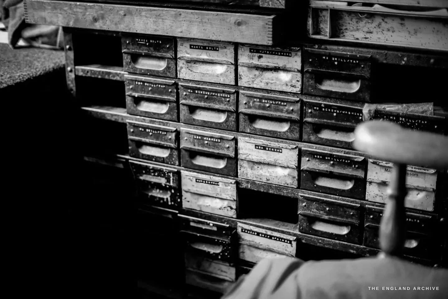 A bank of small metal parts drawers, each hand-labelled - bolts, nuts, washers, drill and tap sizes, springs - a maker’s hand resting at the edge.