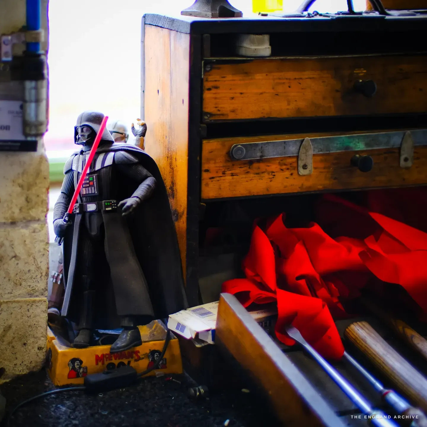 A Darth Vader figurine standing on an old McEwan’s beer crate beside a worn wooden tool chest, a red rag in an open drawer.