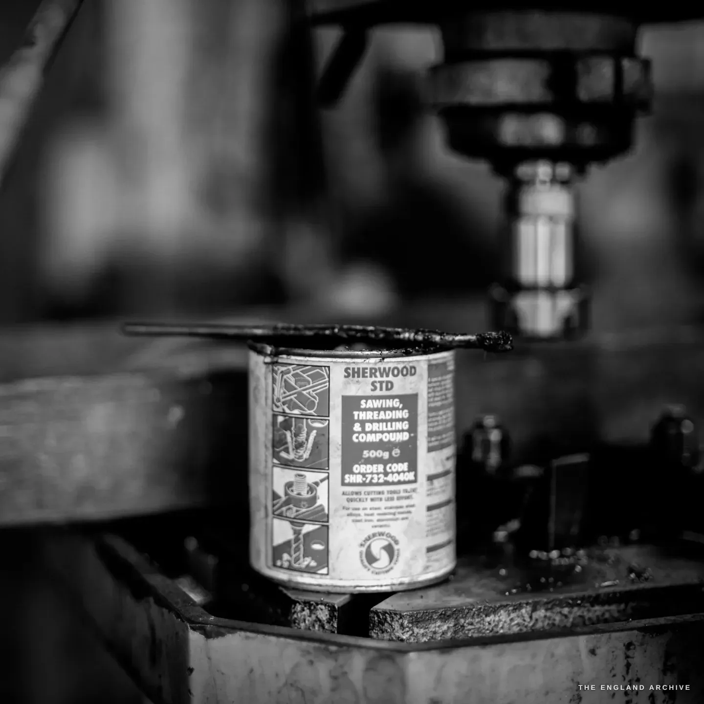 A tin of Sherwood sawing, threading and drilling compound standing on the bed of a pillar drill, the chuck above.
