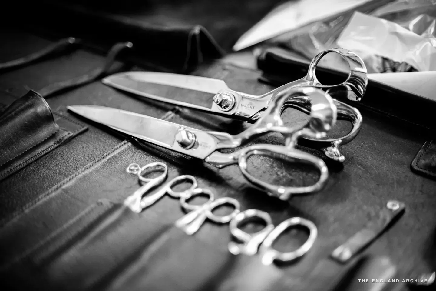 Two finished scissors and a row of small embroidery bows laid out on a dark leather tool roll, the larger shear etched WRIGHT on the blade.
