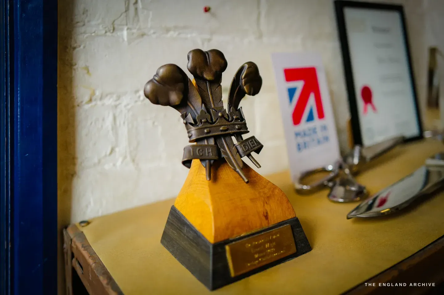 A Prince of Wales feathers trophy with the motto ICH DIEN on a turned wooden plinth, a Made in Britain card and a framed certificate on the shelf beside it.