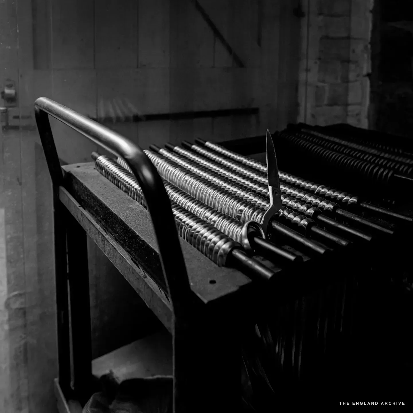 A wheeled trolley stacked with part-finished scissors threaded on rods, one finished blade standing proud in the dark of the shop.