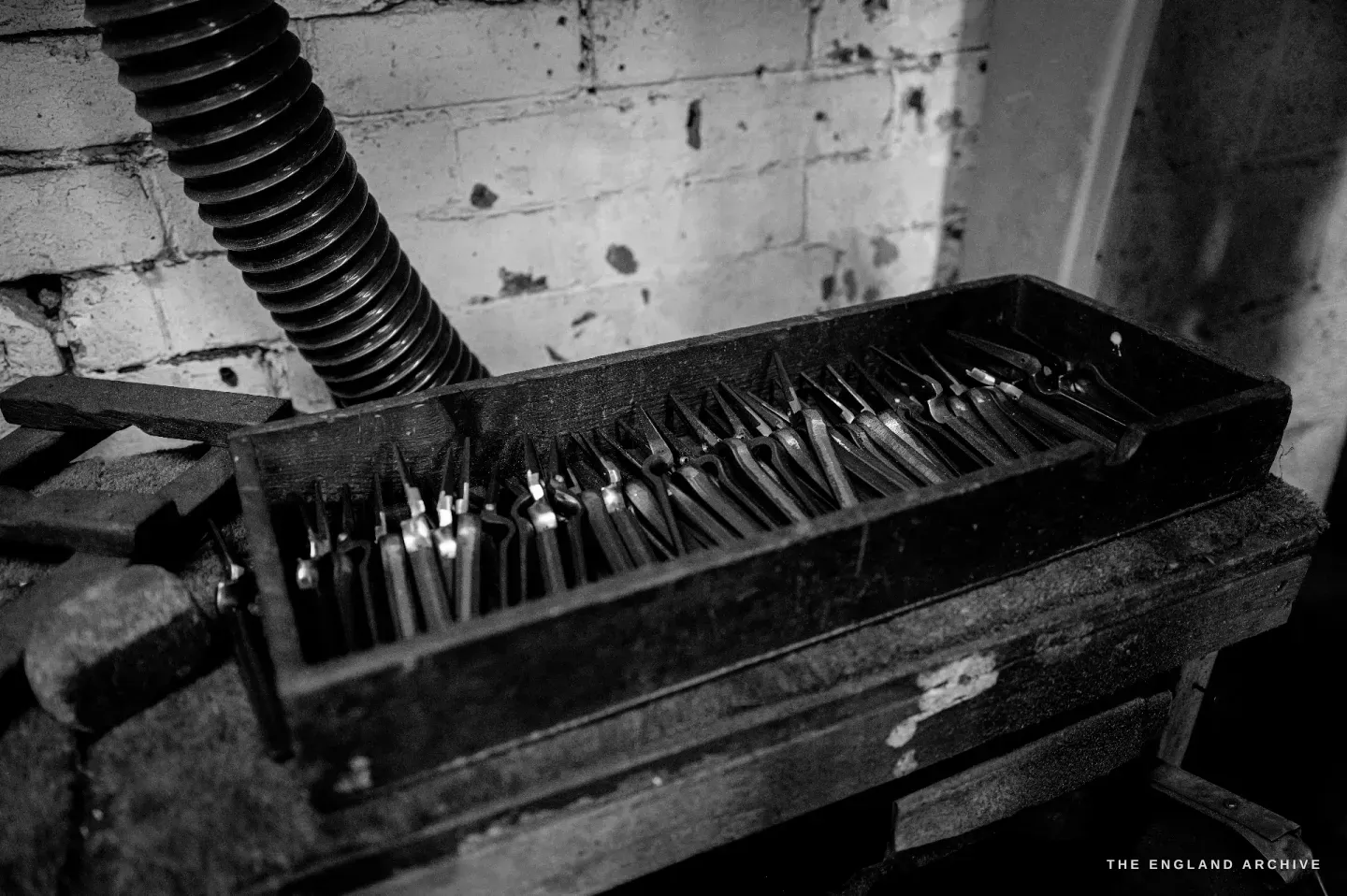 A black tray of part-finished scissor blades standing on edge in rows, an extraction hose curling above.