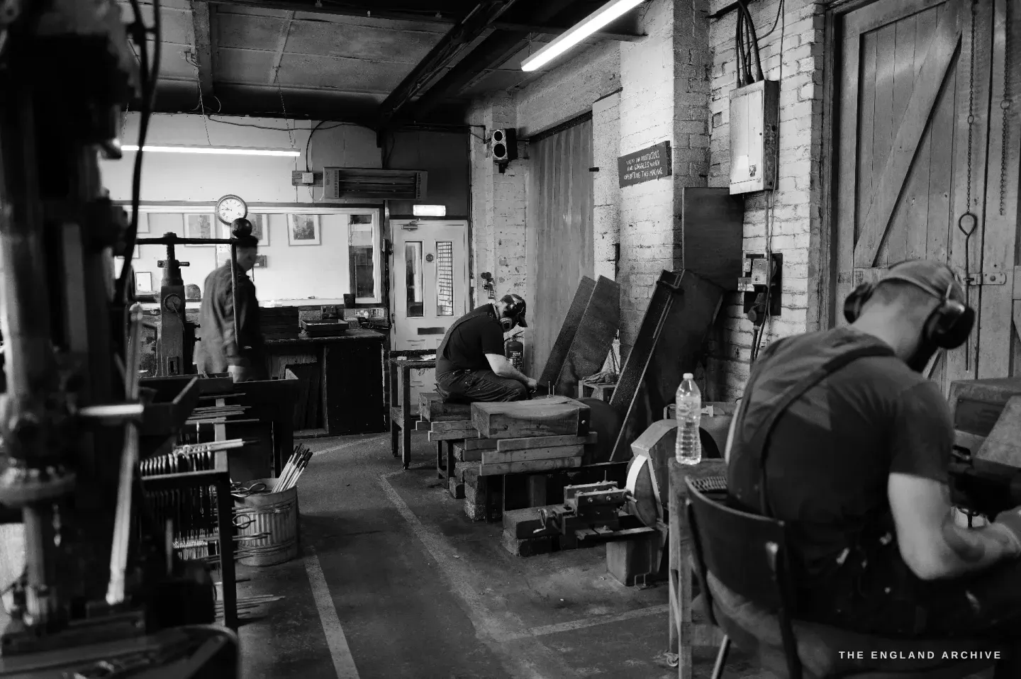 A wide view of the Ernest Wright workshop floor: two makers at their benches, a drill press, the painted safety sign on a brick pillar, daylight from tall windows, a clock on the wall.