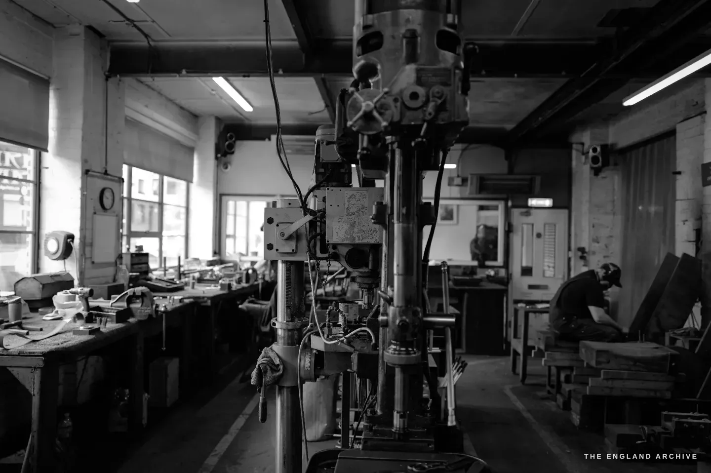 The workshop seen down its length: a tall pillar drill in the foreground, benches along a wall of windows, a maker working at the far end.