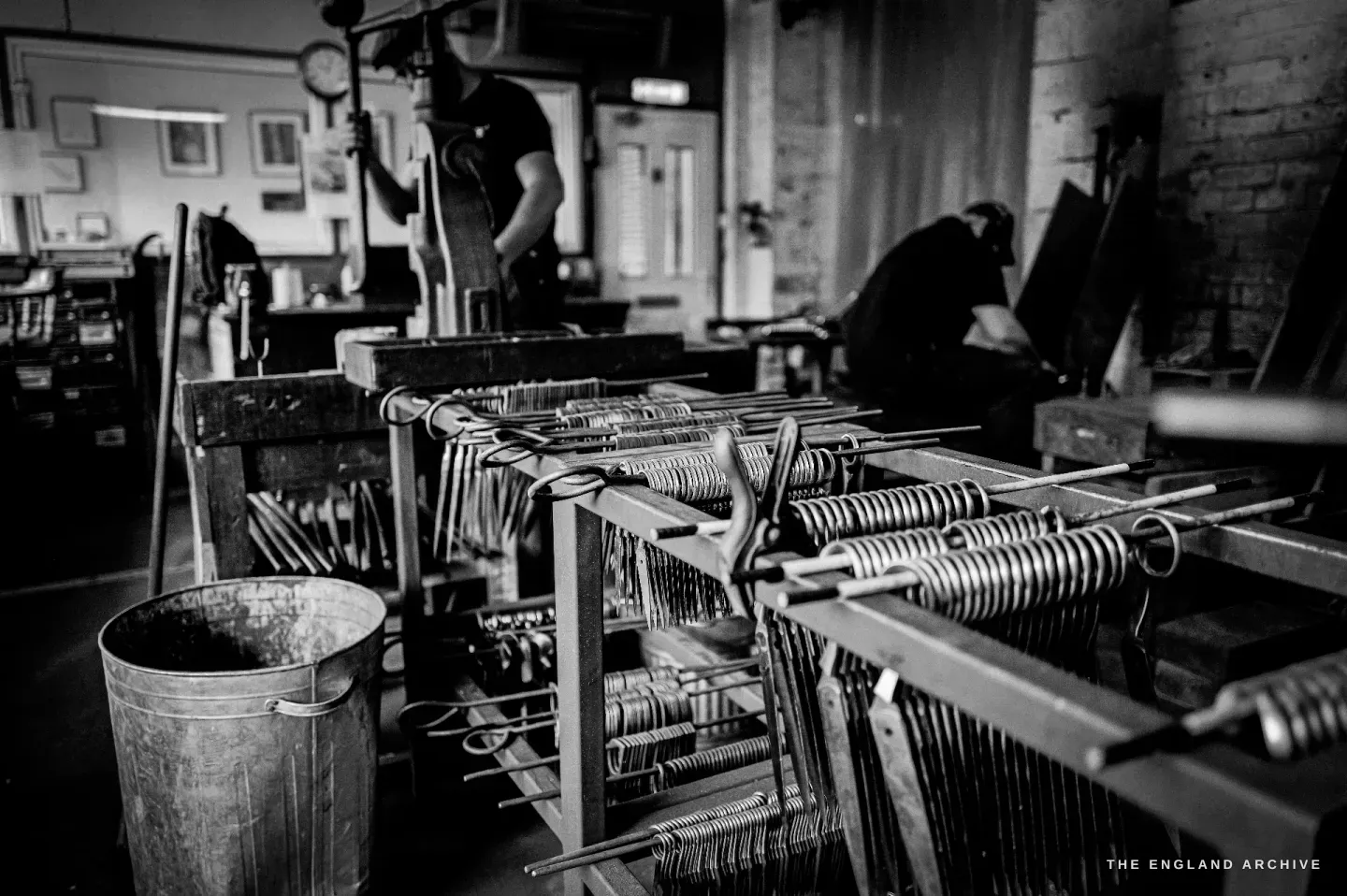 A wide view across the workshop: benches of part-made scissors threaded on rods, a bucket on the floor, two makers working at machines in the background.