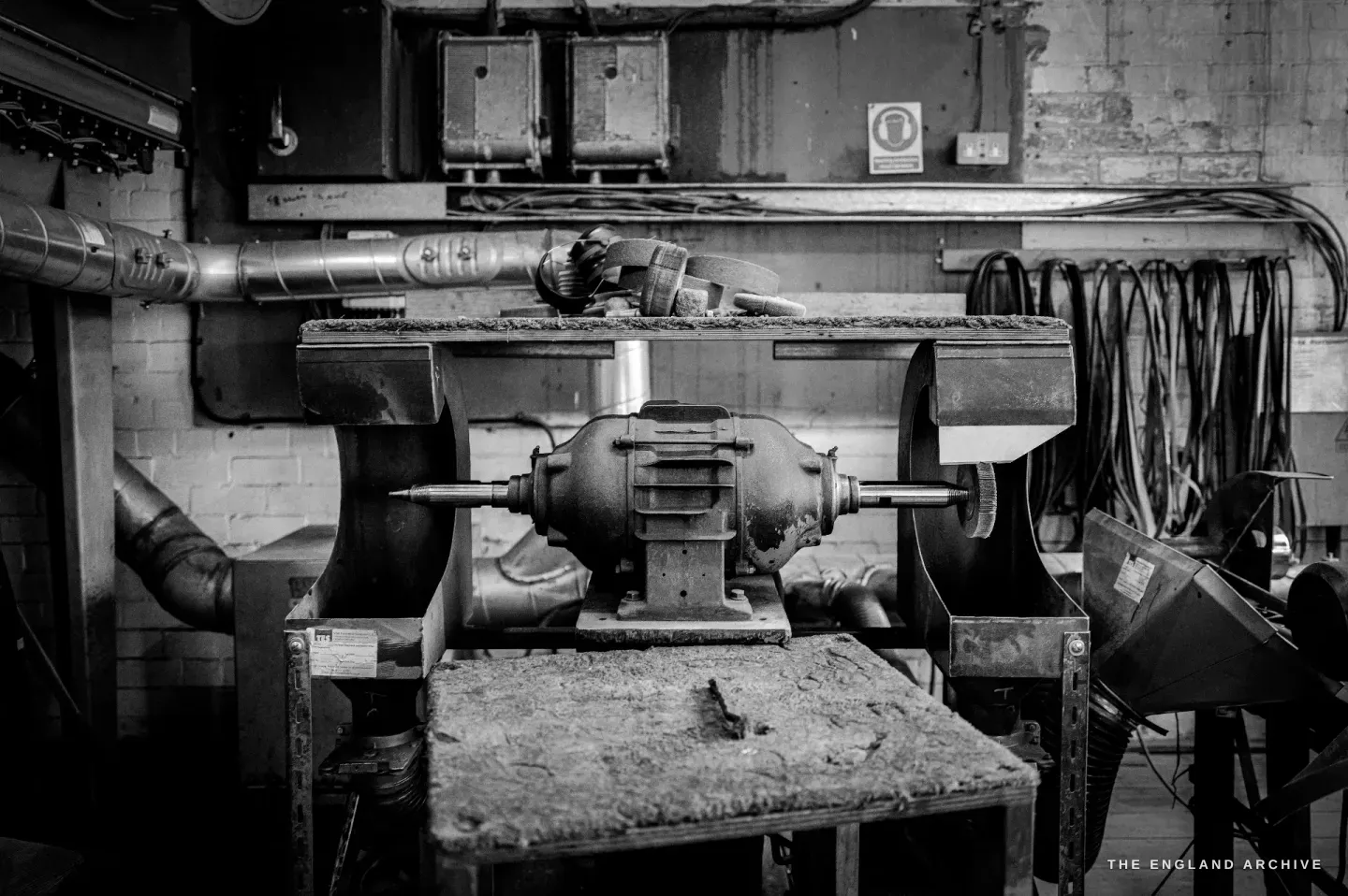 A double-ended grinding and buffing machine on a worn bench, drive shaft and pipework behind it against a whitewashed wall.
