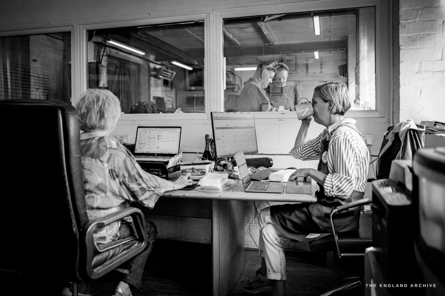 The office: two women at desks with laptops and monitors, one drinking from a mug, and through an internal window two makers at work in the workshop beyond.
