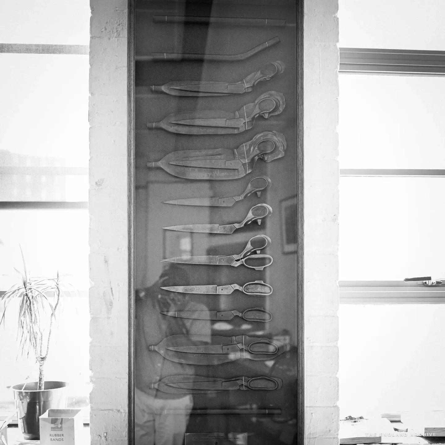 A tall glass display case on the workshop wall holding a column of scissor parts at successive stages of making, from rough blank through to finished blade and bow.